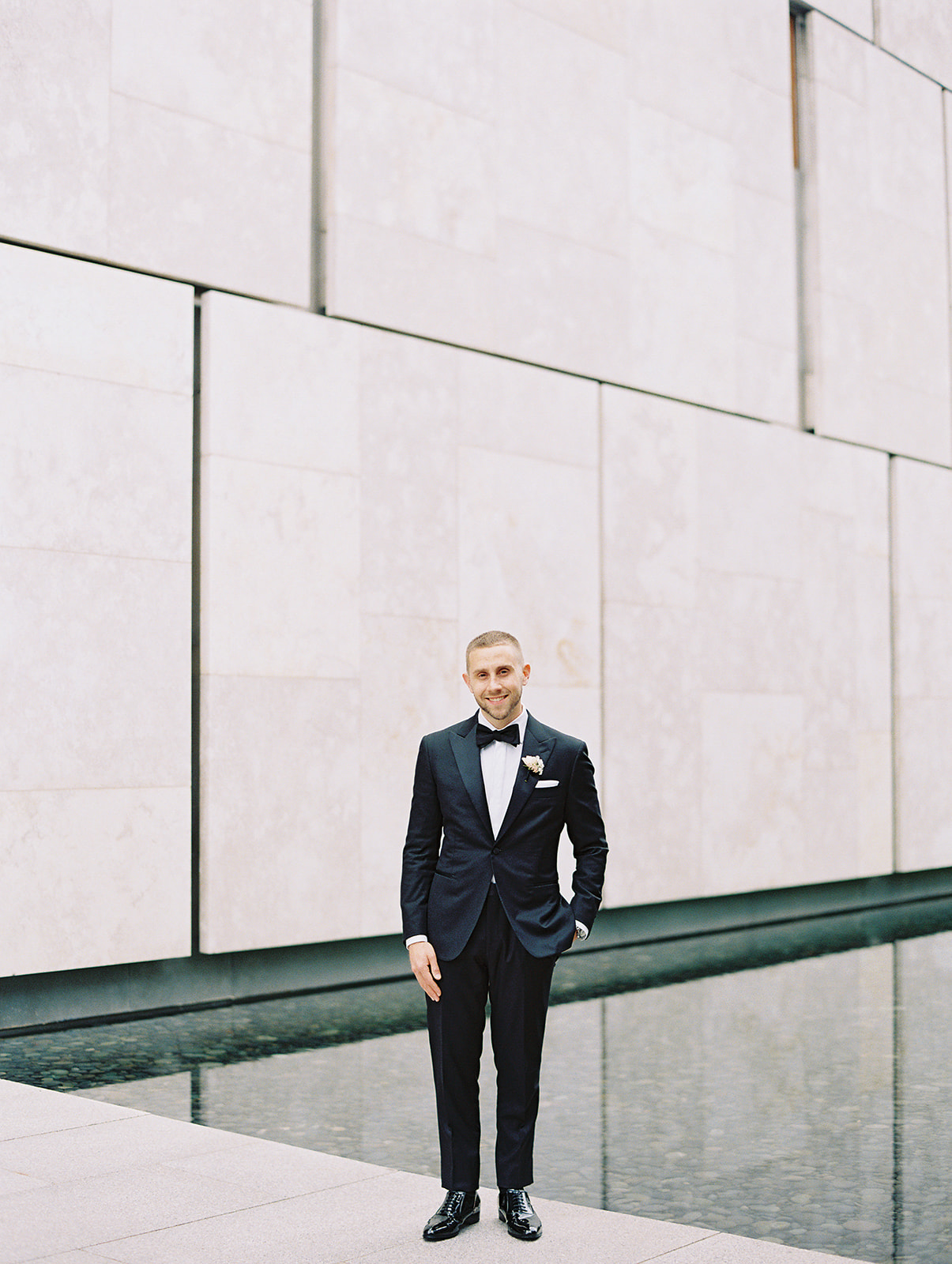 Groom in a black tux against a marble wall and reflecting pool at the Barnes Foundation in Philadelphia