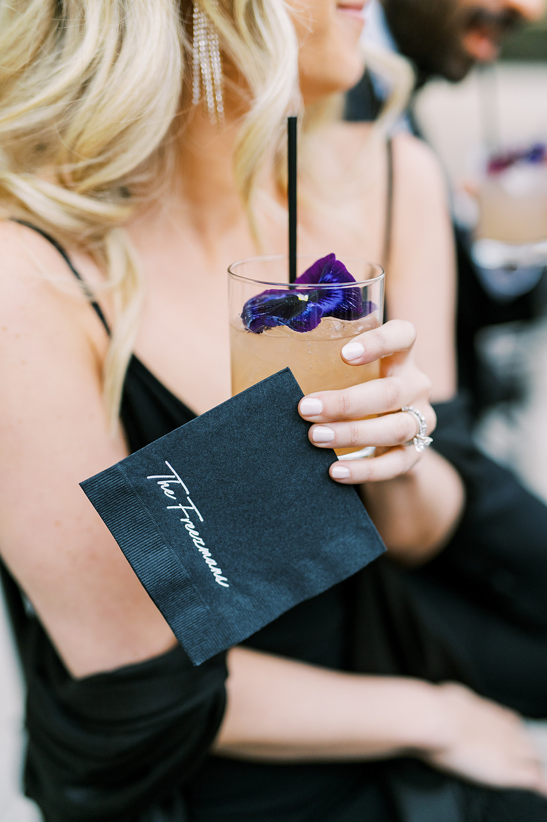 wedding guest holding a signature cocktail beverage with garnish of a purple pansy and a custom black napkin