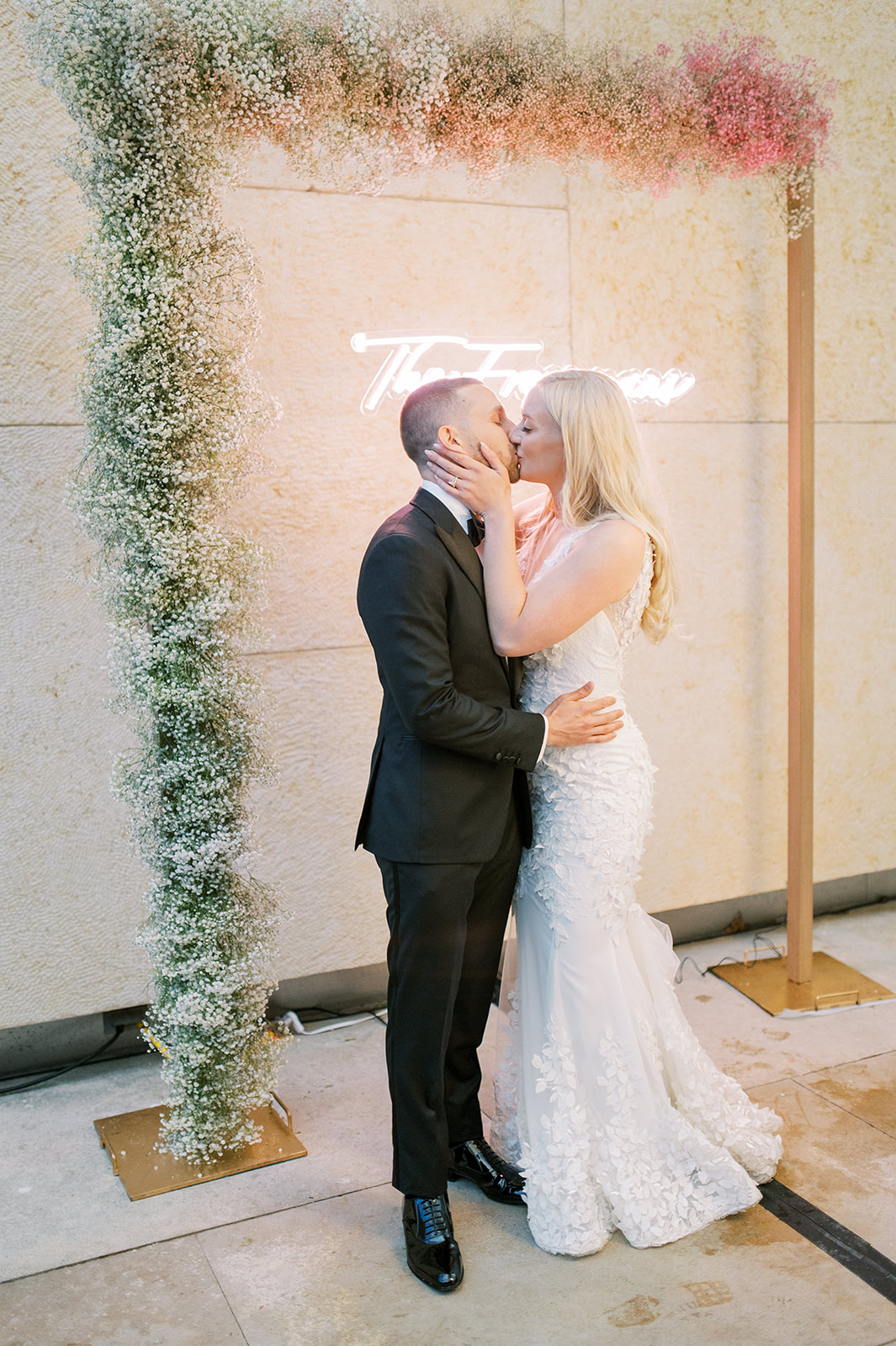 bride and groom kissing under arch of white and pink babies breath in front of neon sign