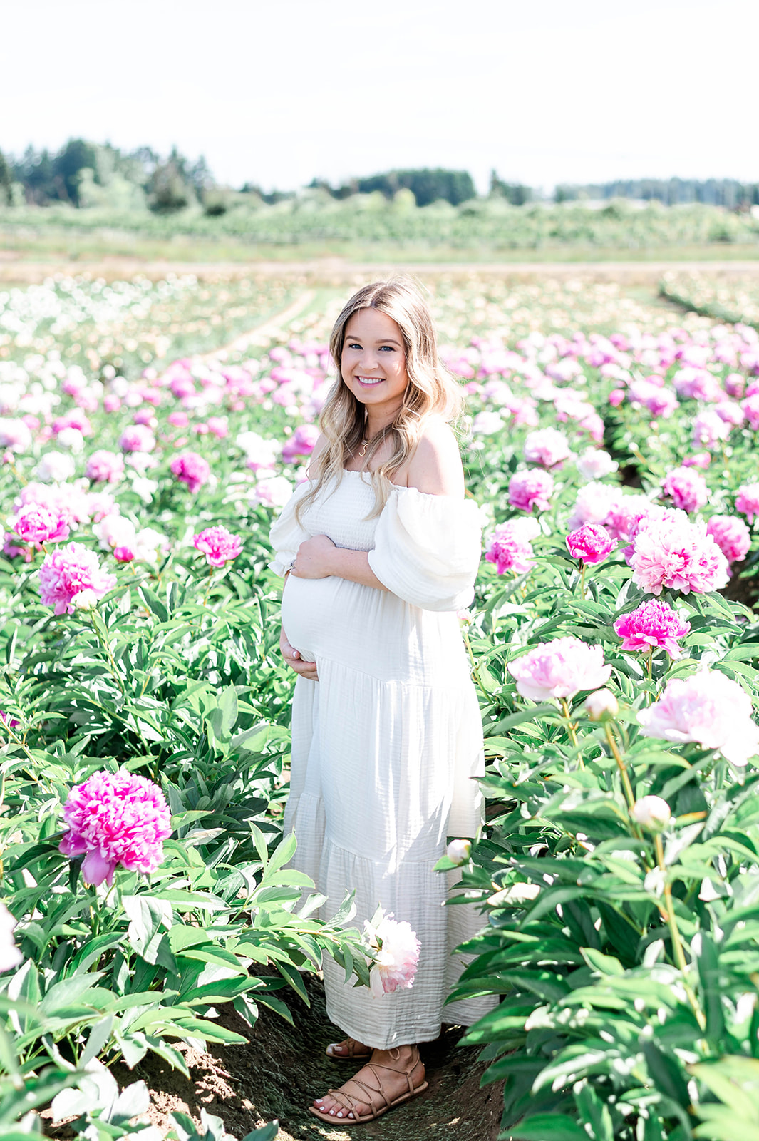 A stunning photograph of a pregnant woman in a white  standing in the midst of a peony flower field 