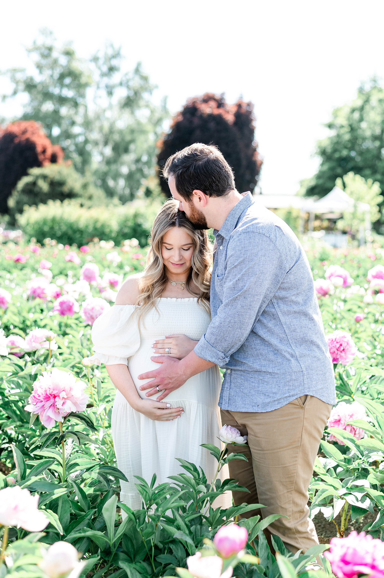 A pregnant woman wearing a flowing white dress with her husband, standing amid a colorful flower field of peonies