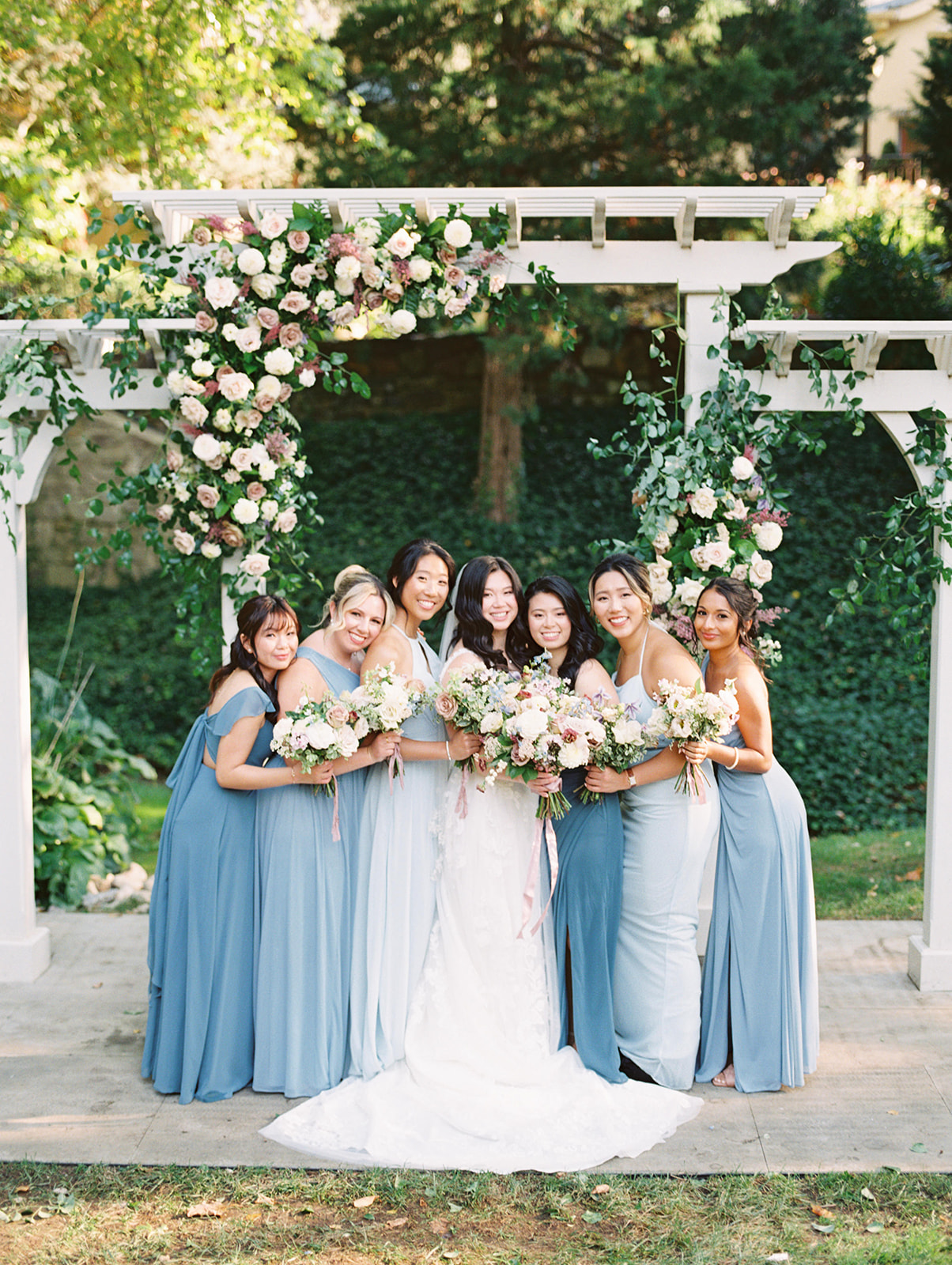 bridesmaids in varying light blue dresses huddle together for a picture under a floral arch at Pomme in Radnor, PA