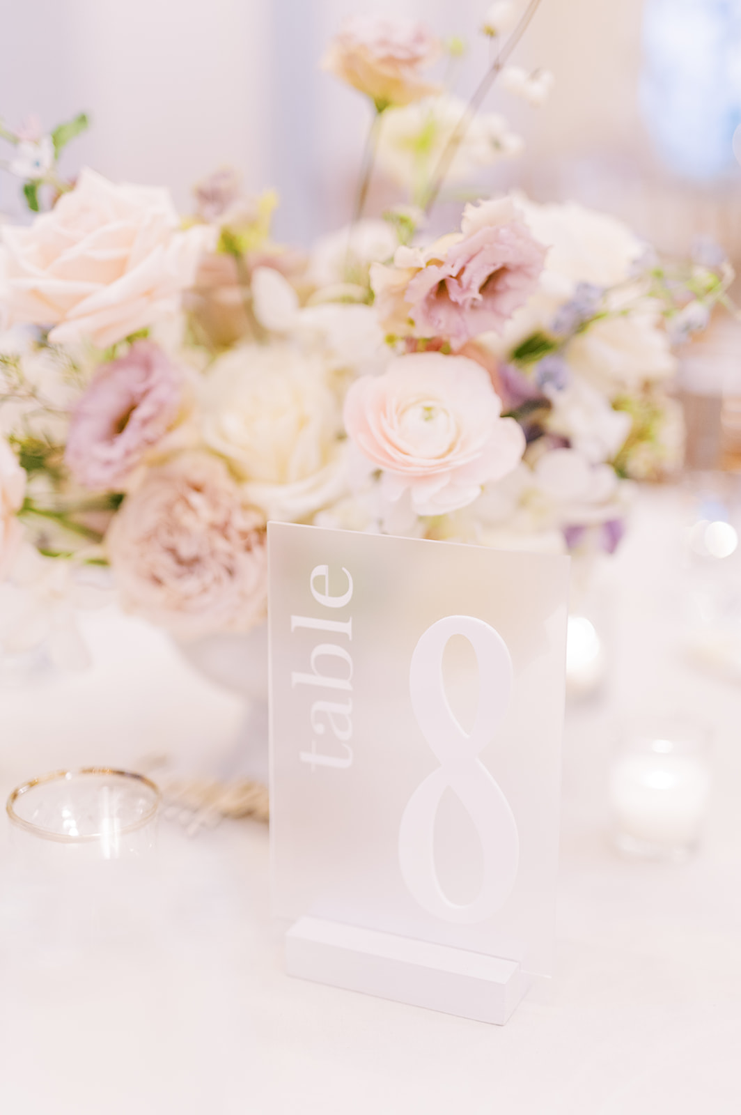 clear frosted acrylic table number with white lettering in front of neutral monochromatic floral centerpiece for wedding