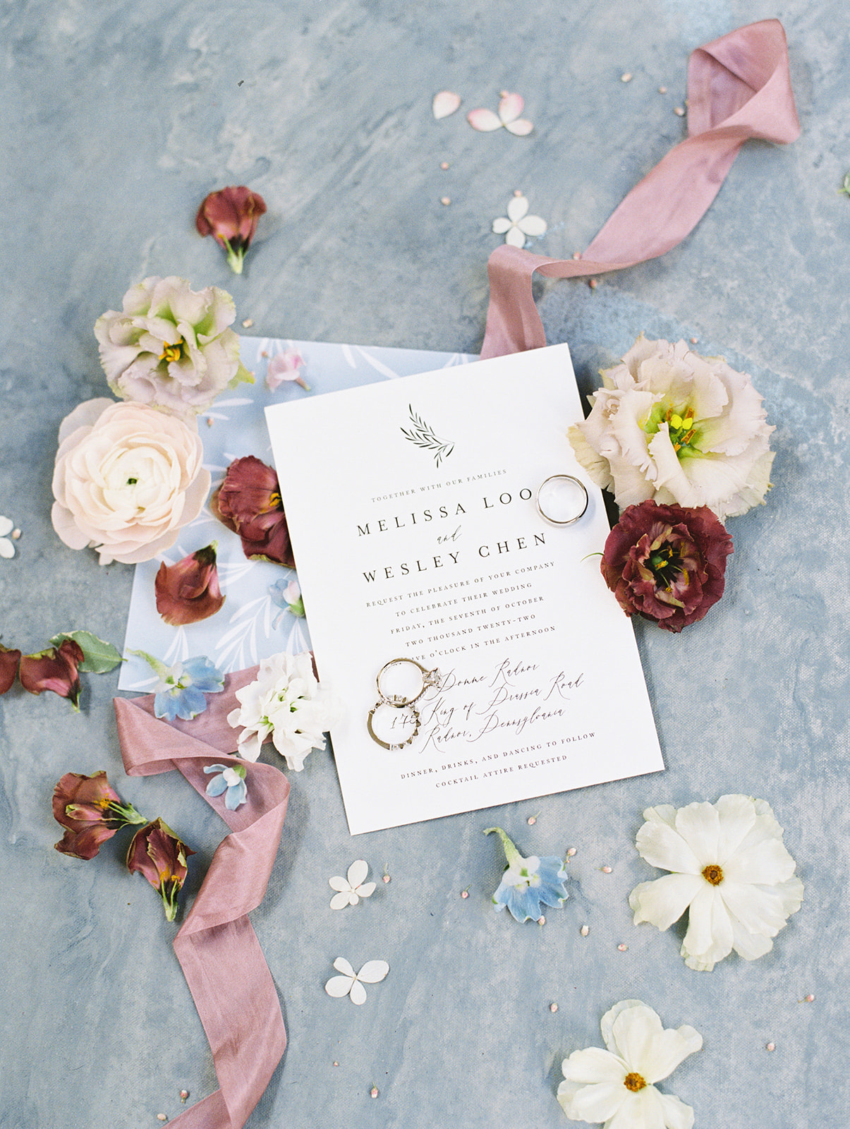 rings sit on invitation with white and burgundy flowers on painted french blue surface
