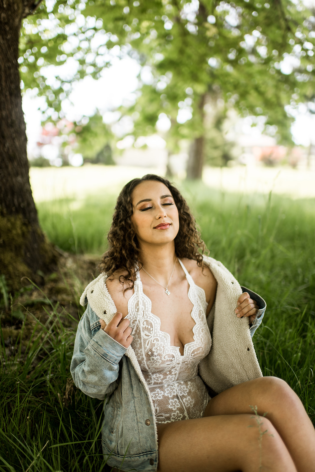 woman with curly hair wears a jean jacket and white lingerie while sitting under a tree for her boudoir session outdoors