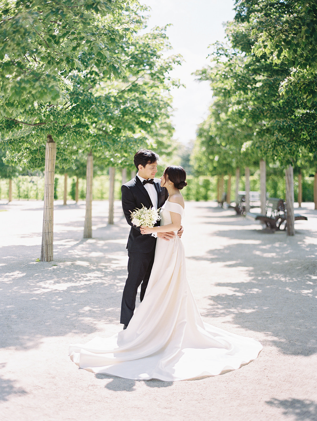 bride and groom embrace on sunny wedding day surrounded by trees at Longwood Gardens in Kennet Square PA