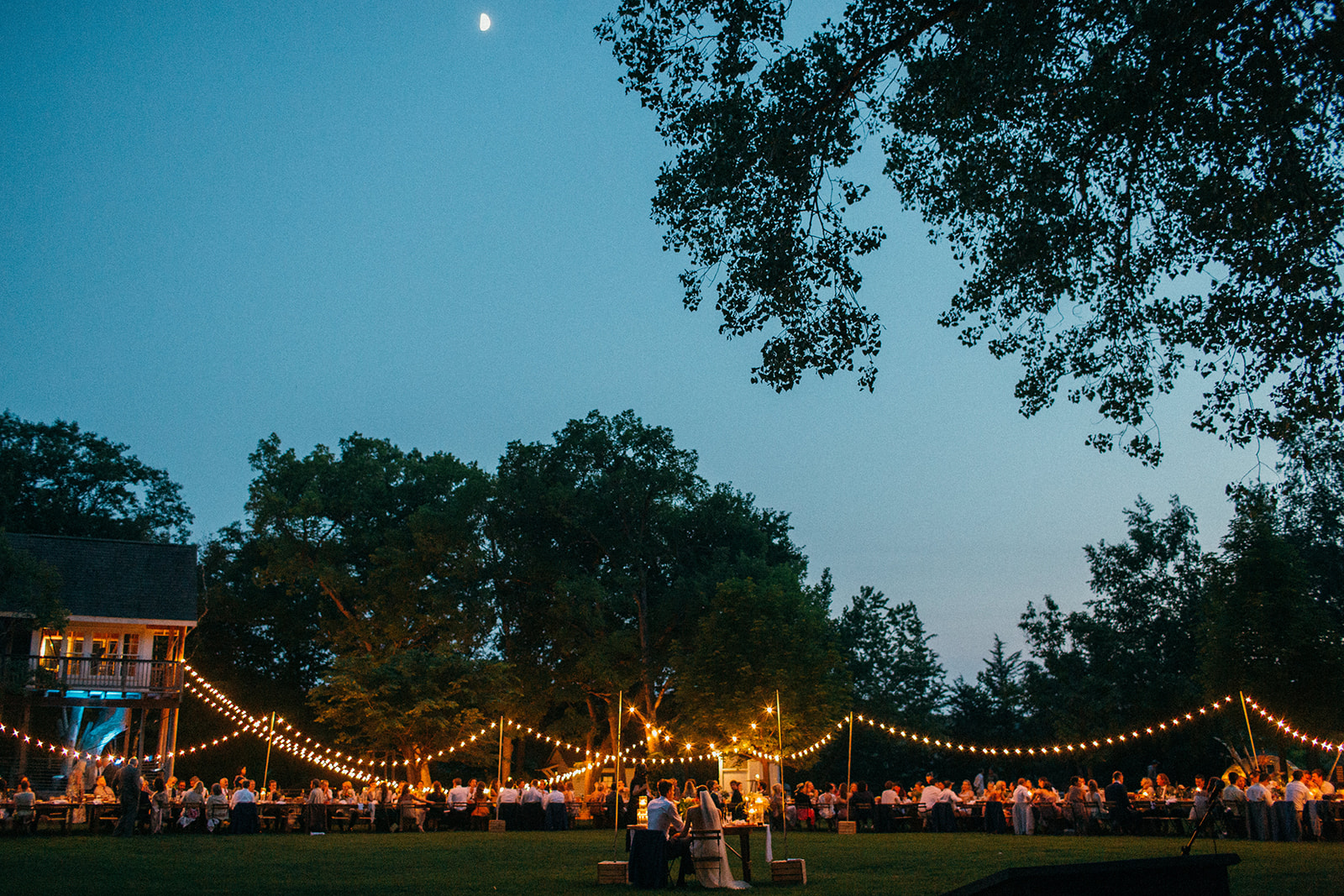 Wedding couple sits down surrounded by their guests under a moonlit sky and string lights at Camp Wandawega