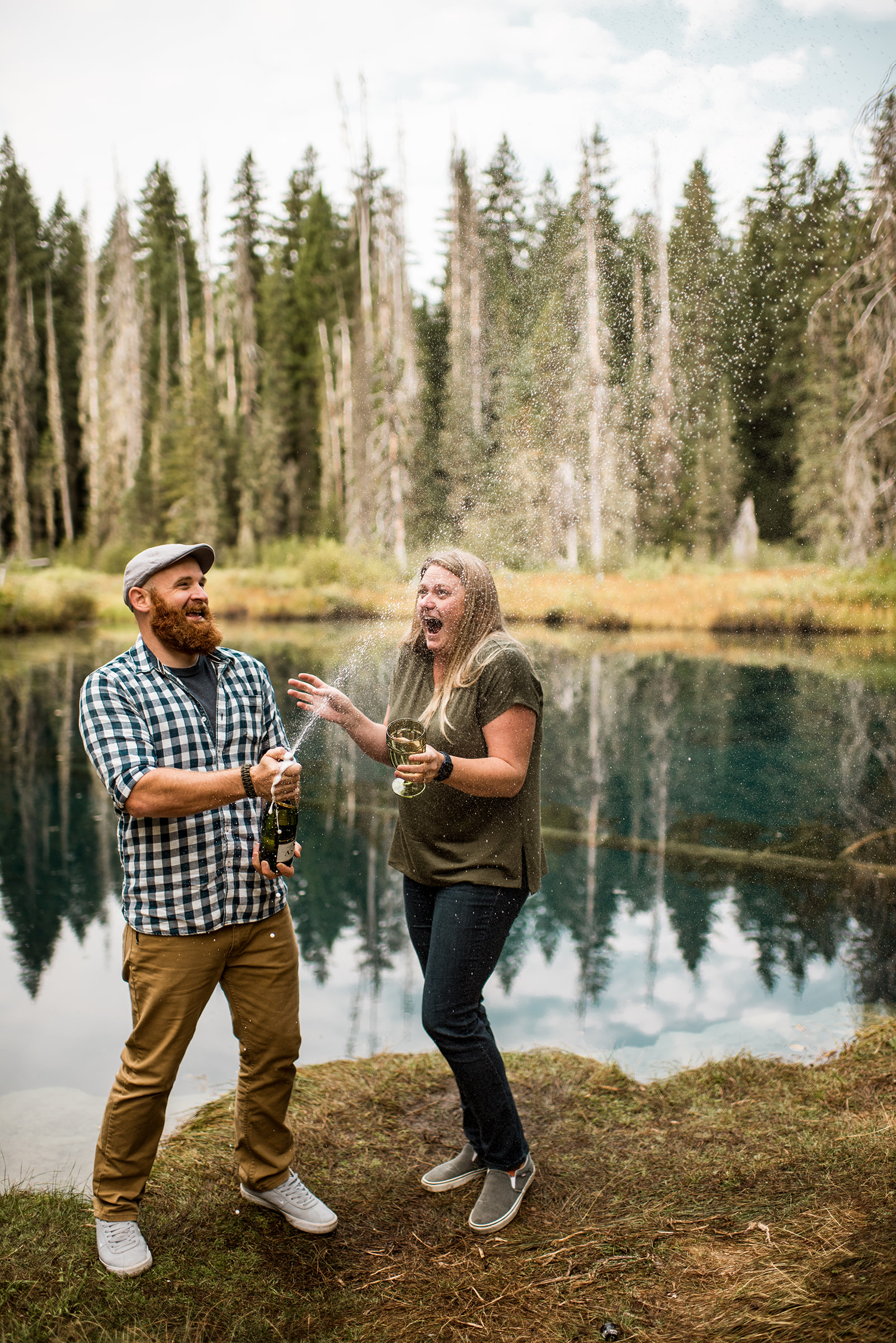 engaged couple pops champagne bottle in front of lake in Mt. Hood Wilderness