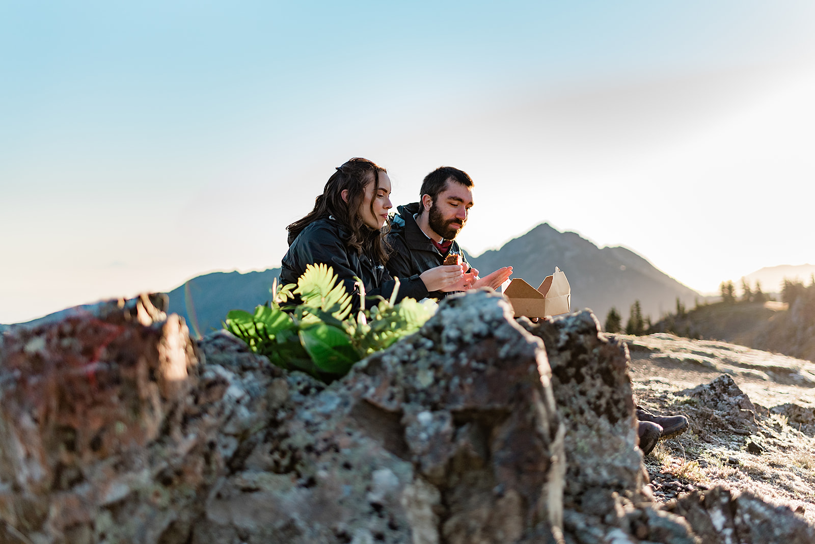 This is a picture of a bride and groom during their elopement they are eating a snack on a mountain.