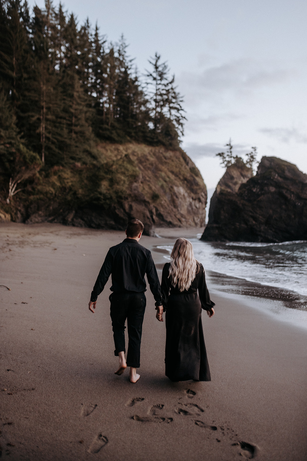 A barefoot eloping couple wearing all black walking away at Secret Beach in Oregon.