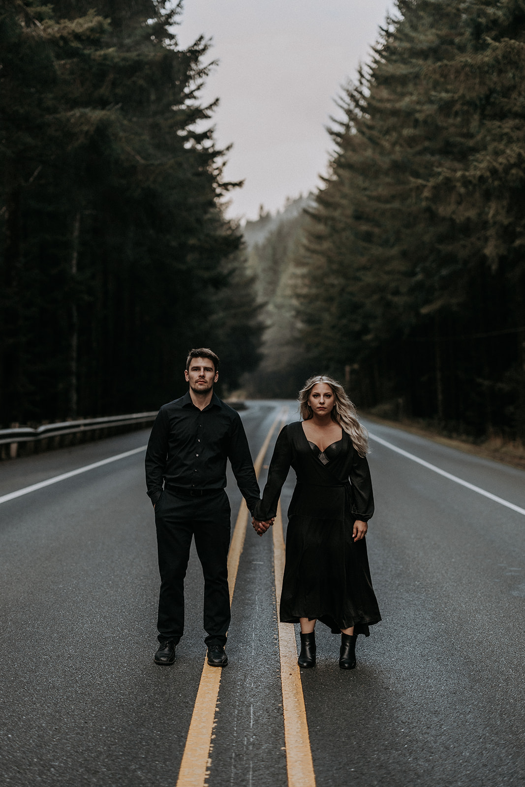 An elopement couple wearing all black with serious faces holding hands on a road in Southern Oregon with pine trees.