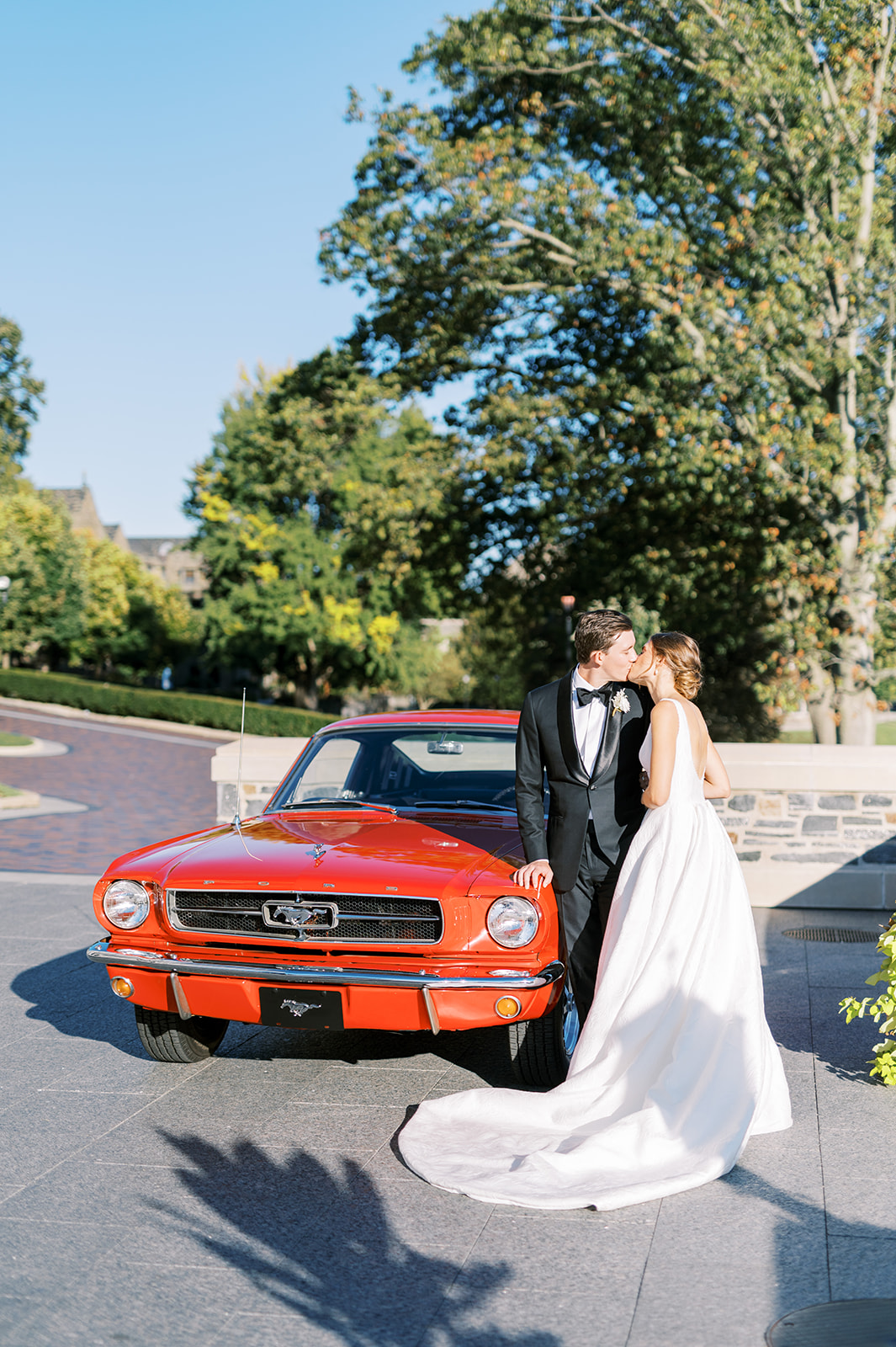 bride and groom kiss in front of classic red mustang for sunny wedding day at villanova