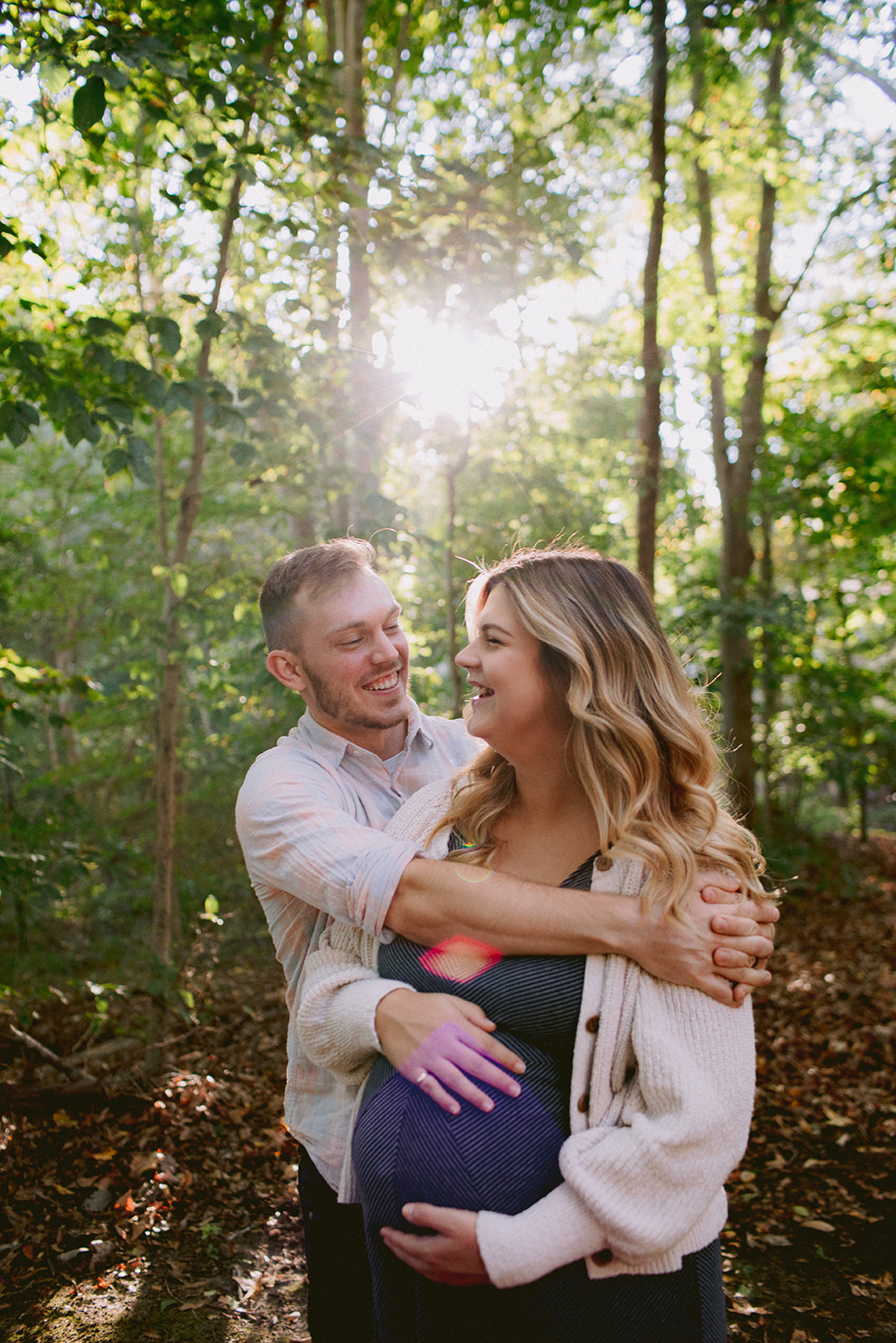 A preganant couple laughing in the woods near their home.