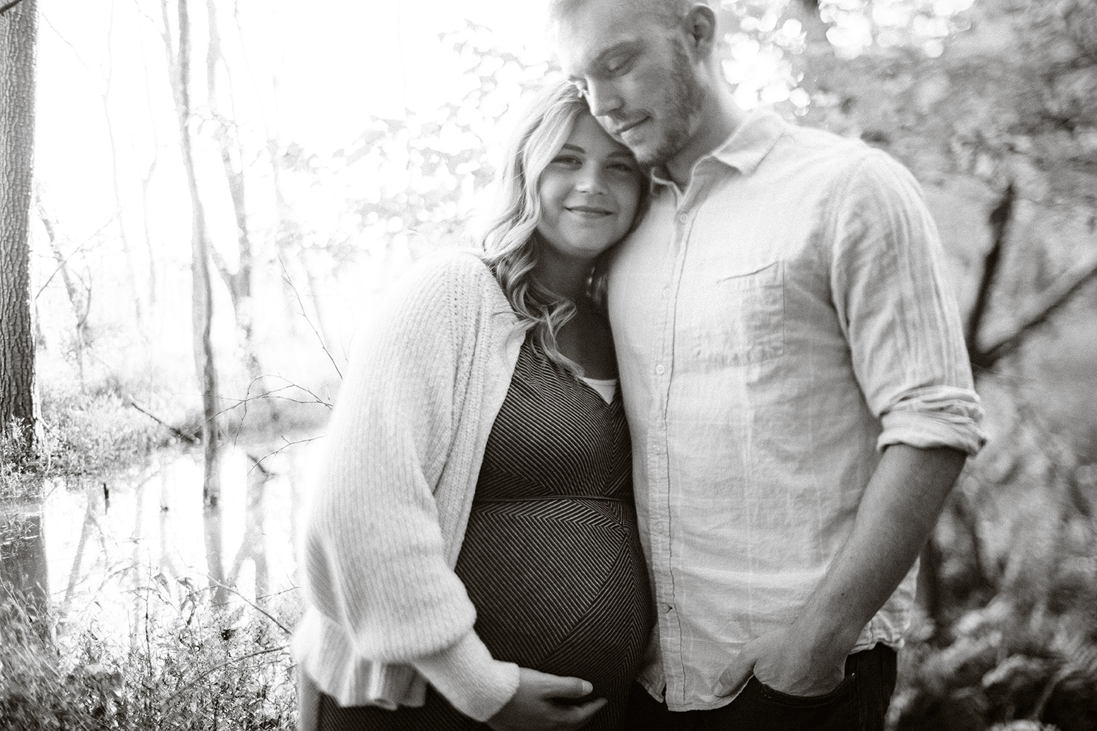 A pregnant couple hug in the woods.
