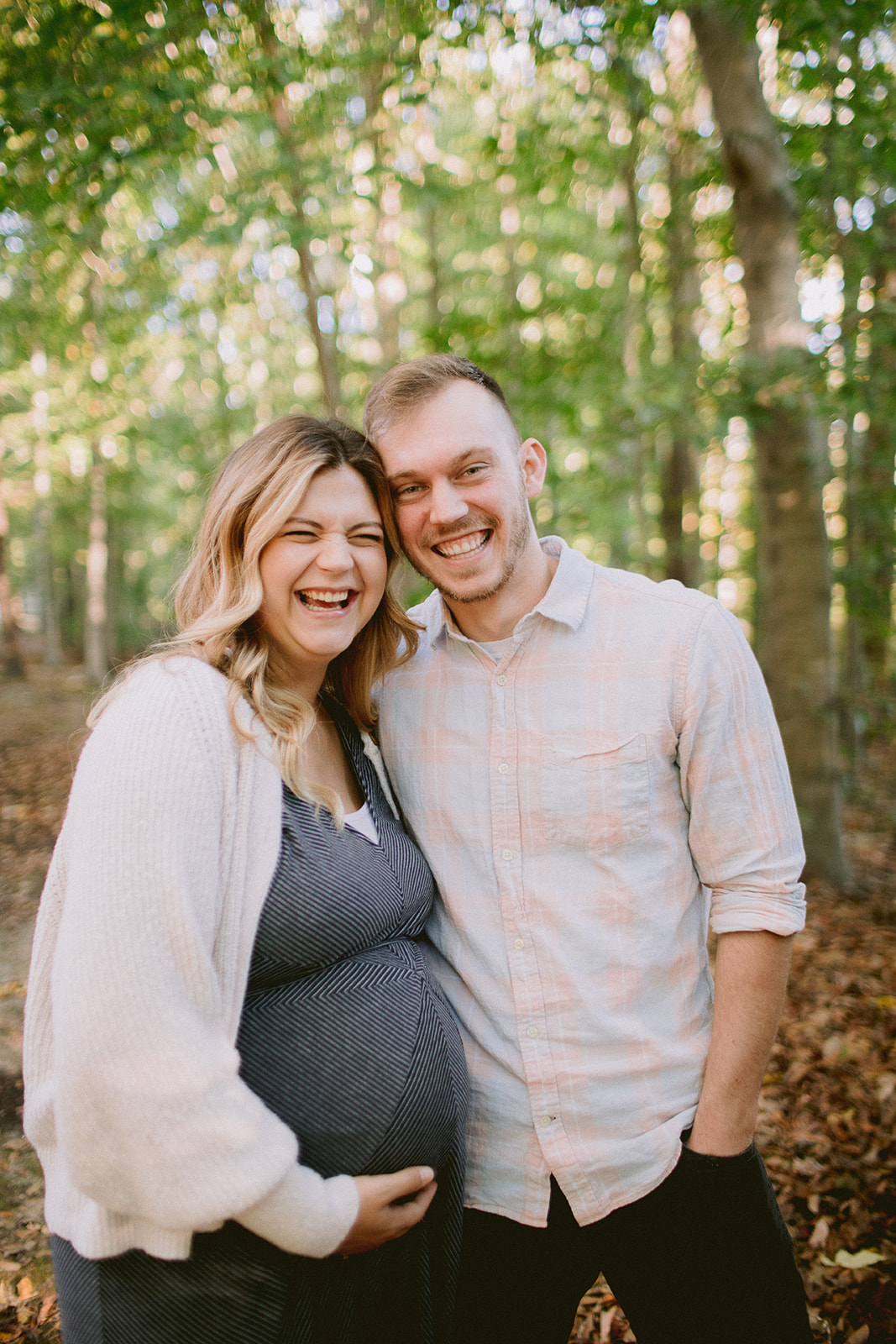 A preganant couple laughing in the woods near their home.
