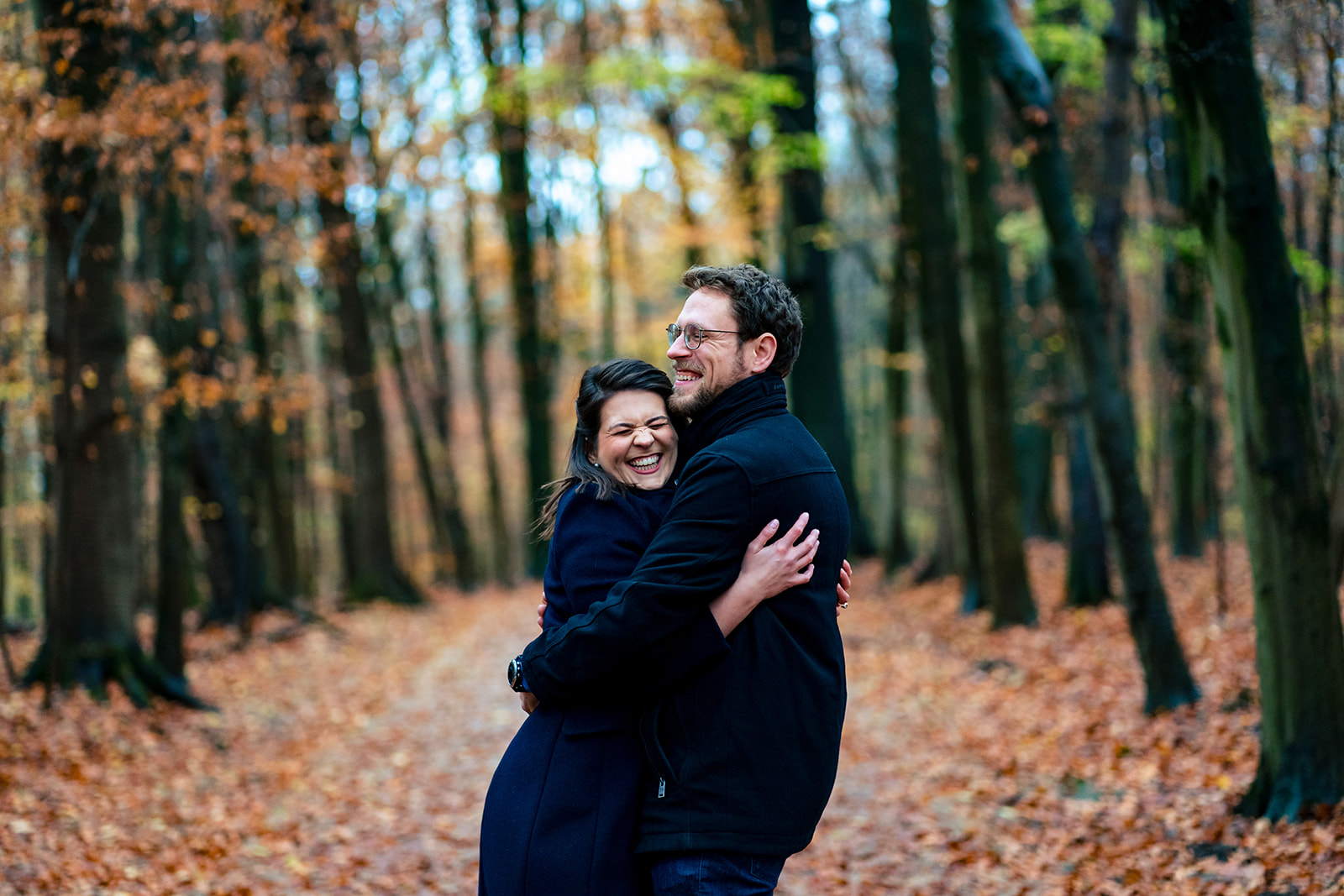 Un couple d'amoureux s'enlacent en riant sur un chemin pleins de feuilles au milieu du bois de la Cambre à Bruxelles