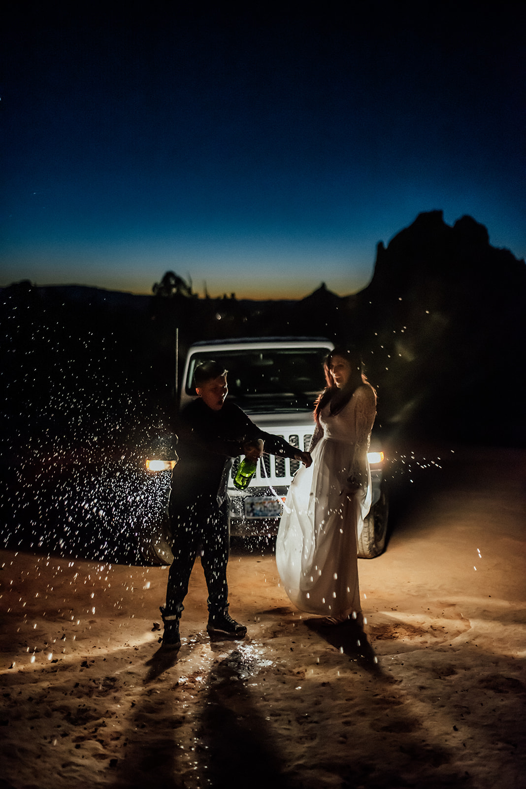 lbtgqia+ couple spraying champagne in front of a jeep with its lights on and sunset behind.