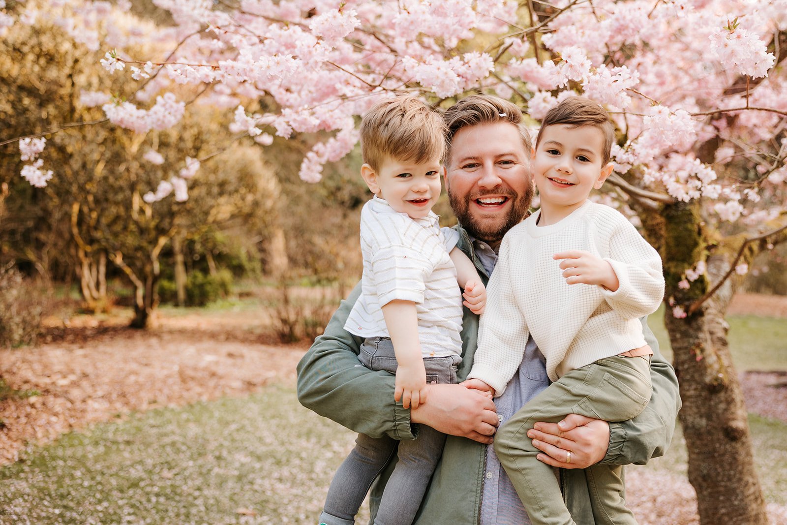 A close-up photo of a smiling father carrying his two young sons in front of a cherry blossom tree. 