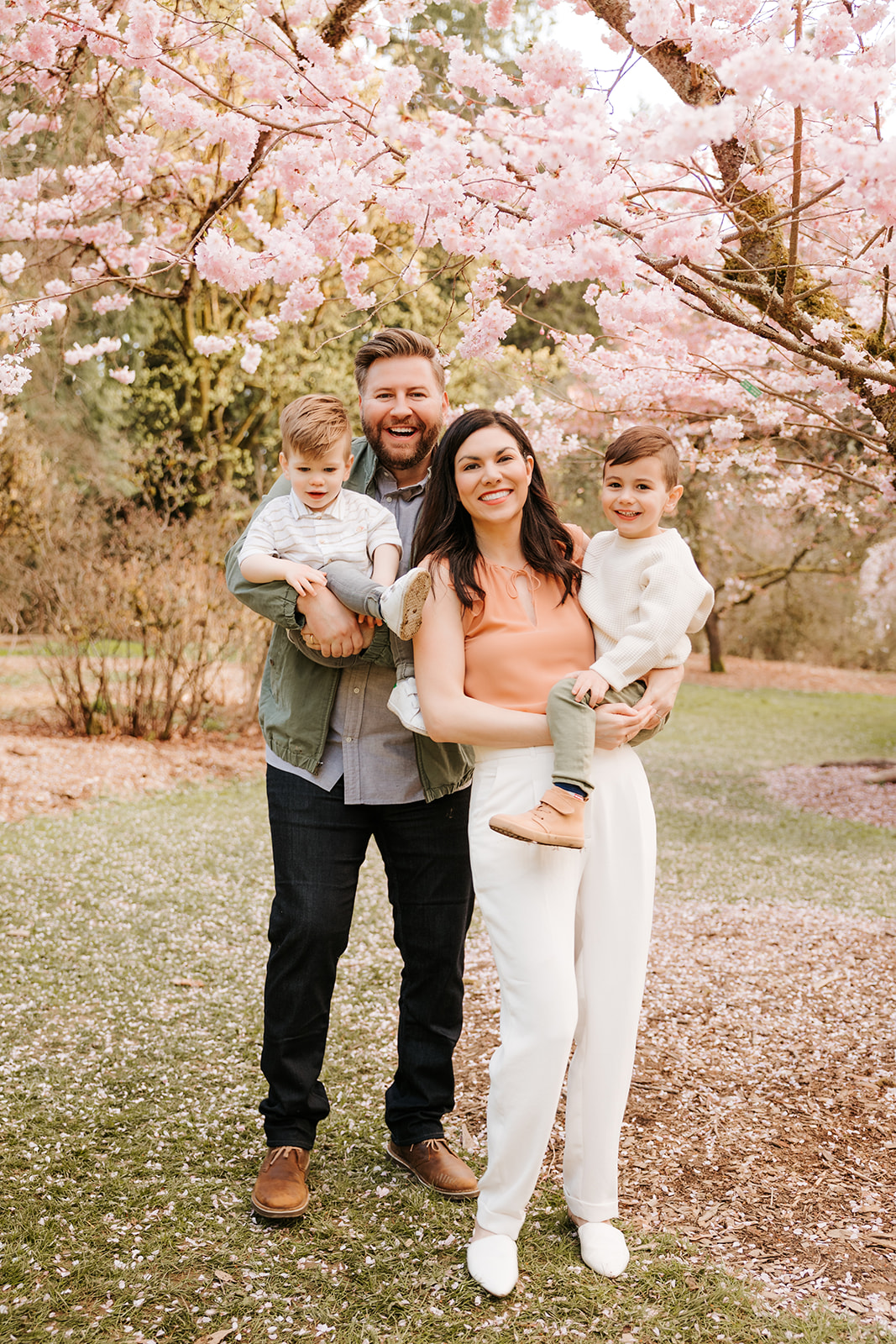 A portrait photo of a family of four standing together in front of a cherry blossom tree. 