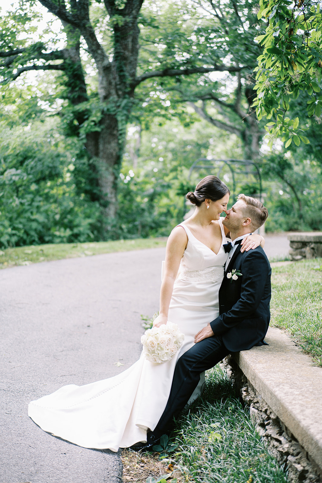 The bride sits on the groom's lap on the stone wall in front of the mansion at Burritt on the Mountain
