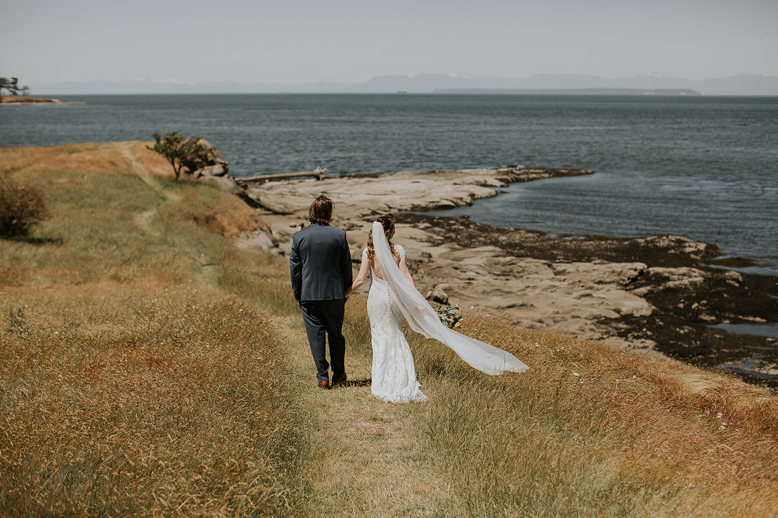 A bride and groom walk away down a path through a field holding hands 