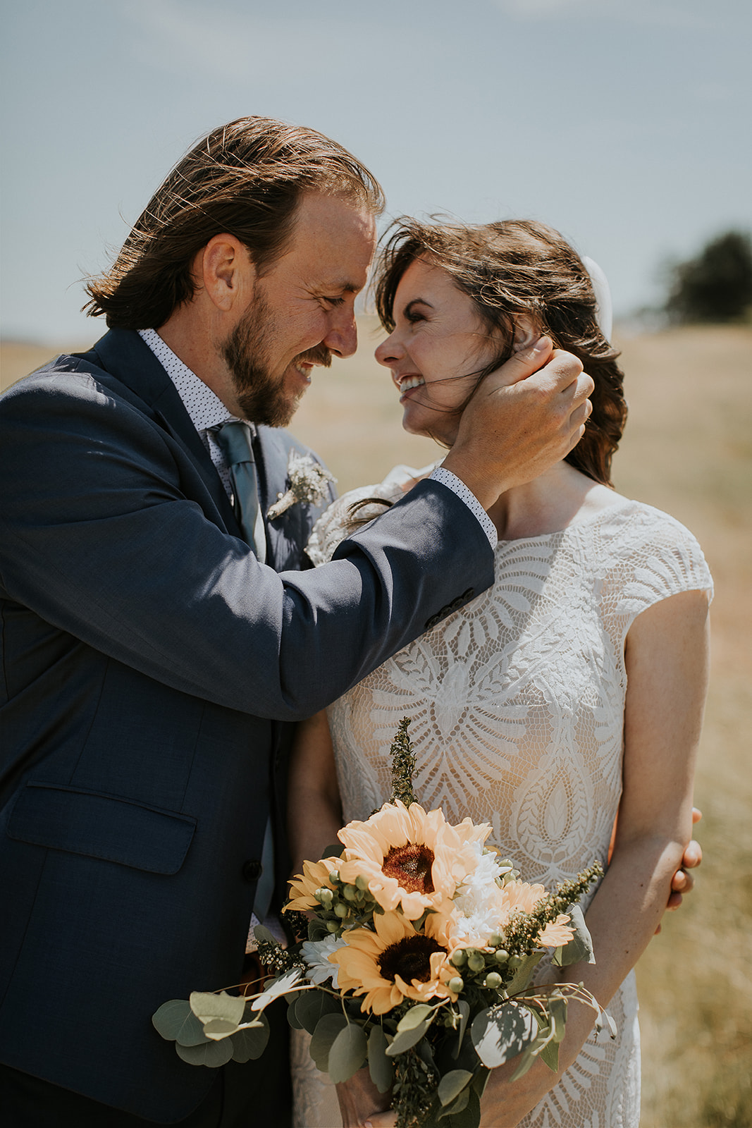 A groom in a blue suit touches his brides hair while she holds sunflowers 