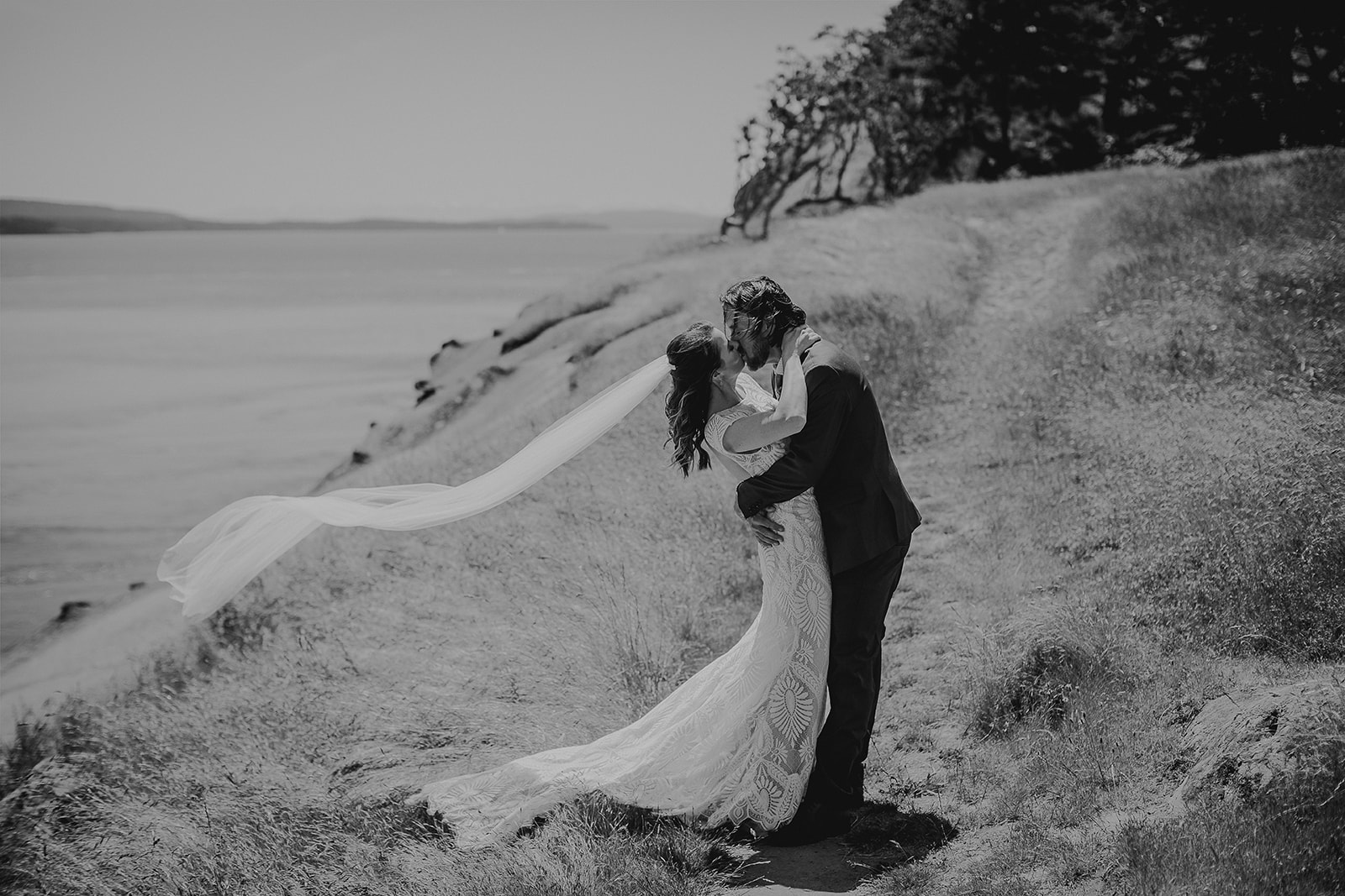 a groom give his bride a big kiss and leans her back while her veil blows in the wind