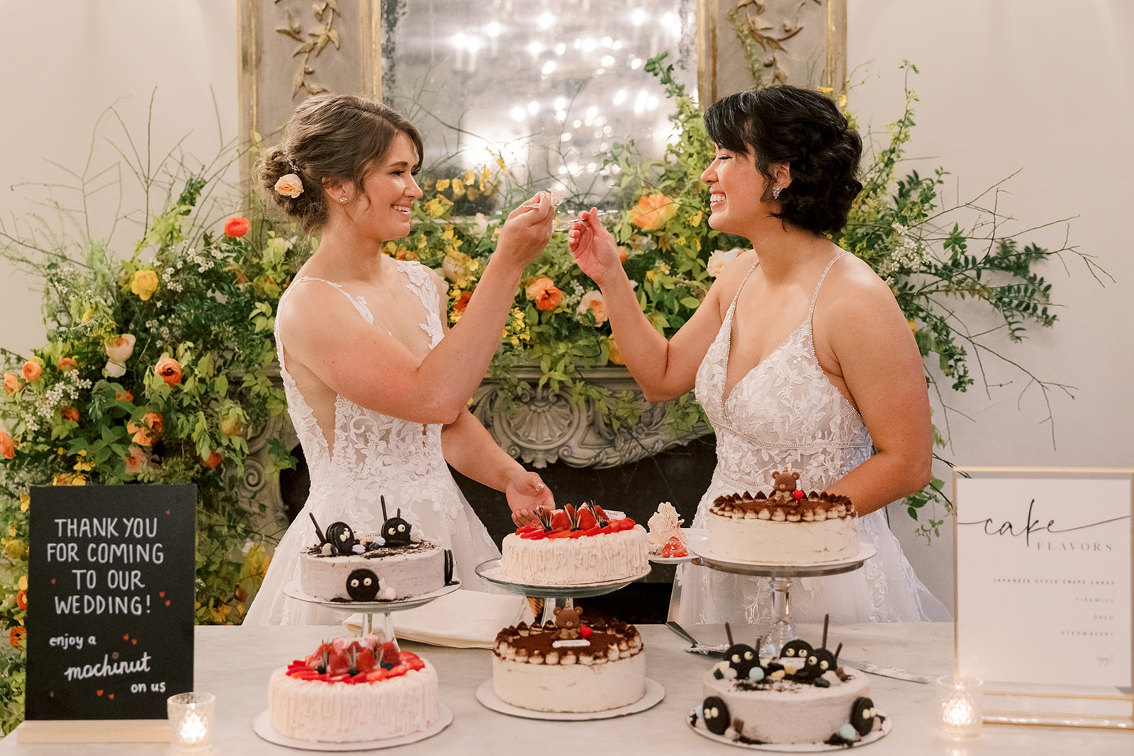 two brides stand behind a cake table swapping forks of cake