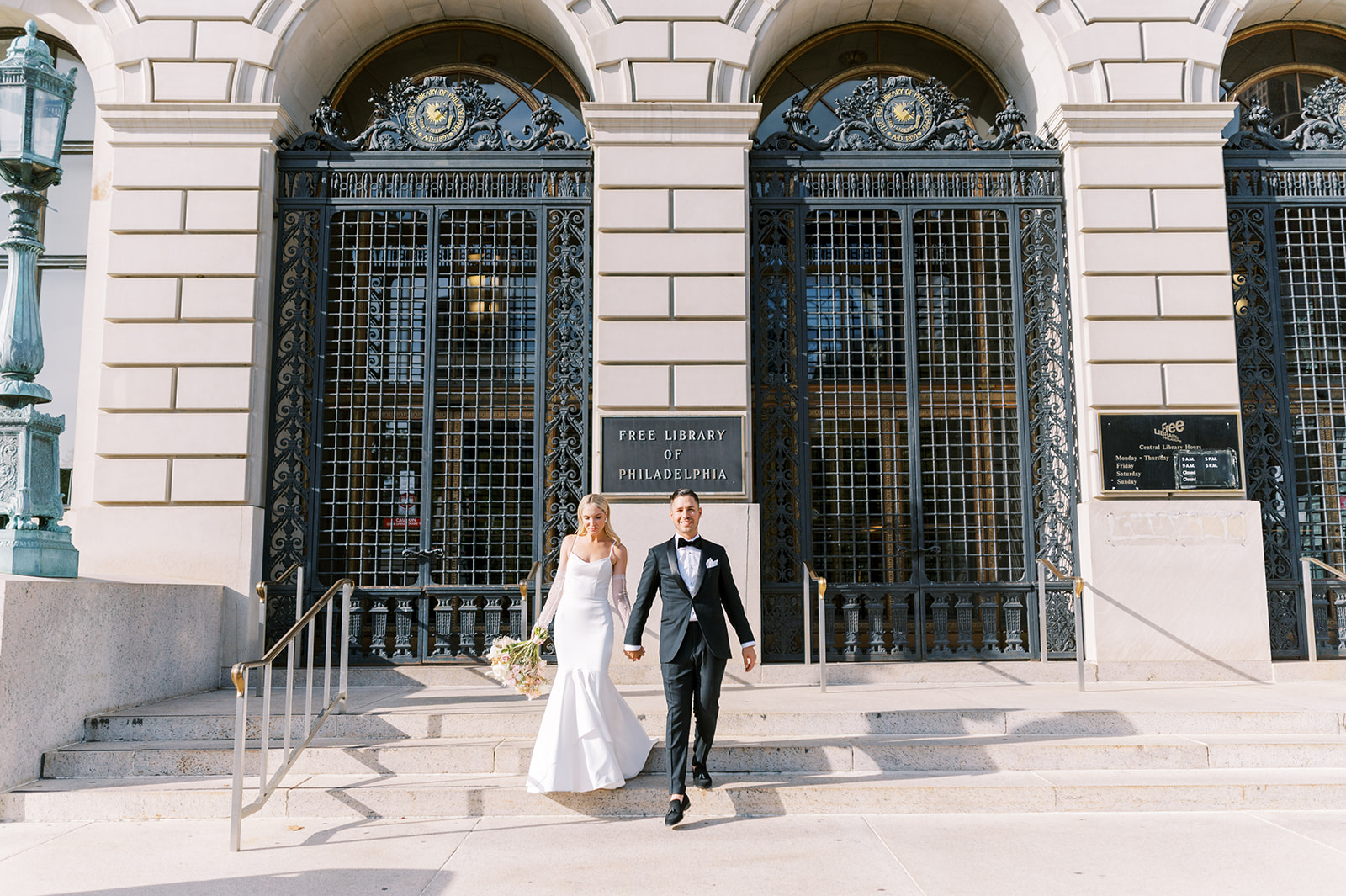 chic bride and groom hold hands as they walk down the Free Library of Philadelphia on their sunny wedding day