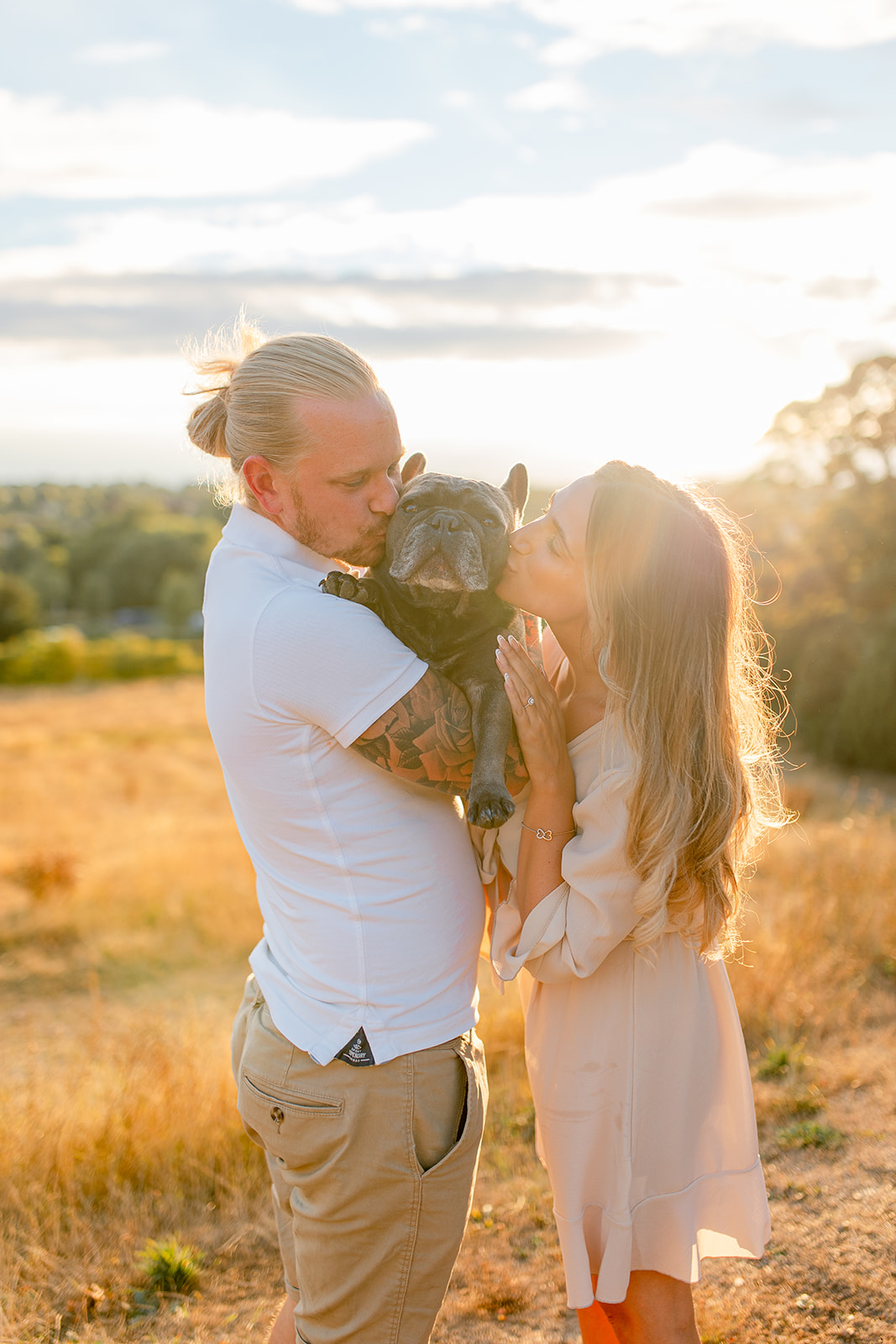 A couple standing in the gorgeous sunlight, side by side holding and kissing their French bulldog.