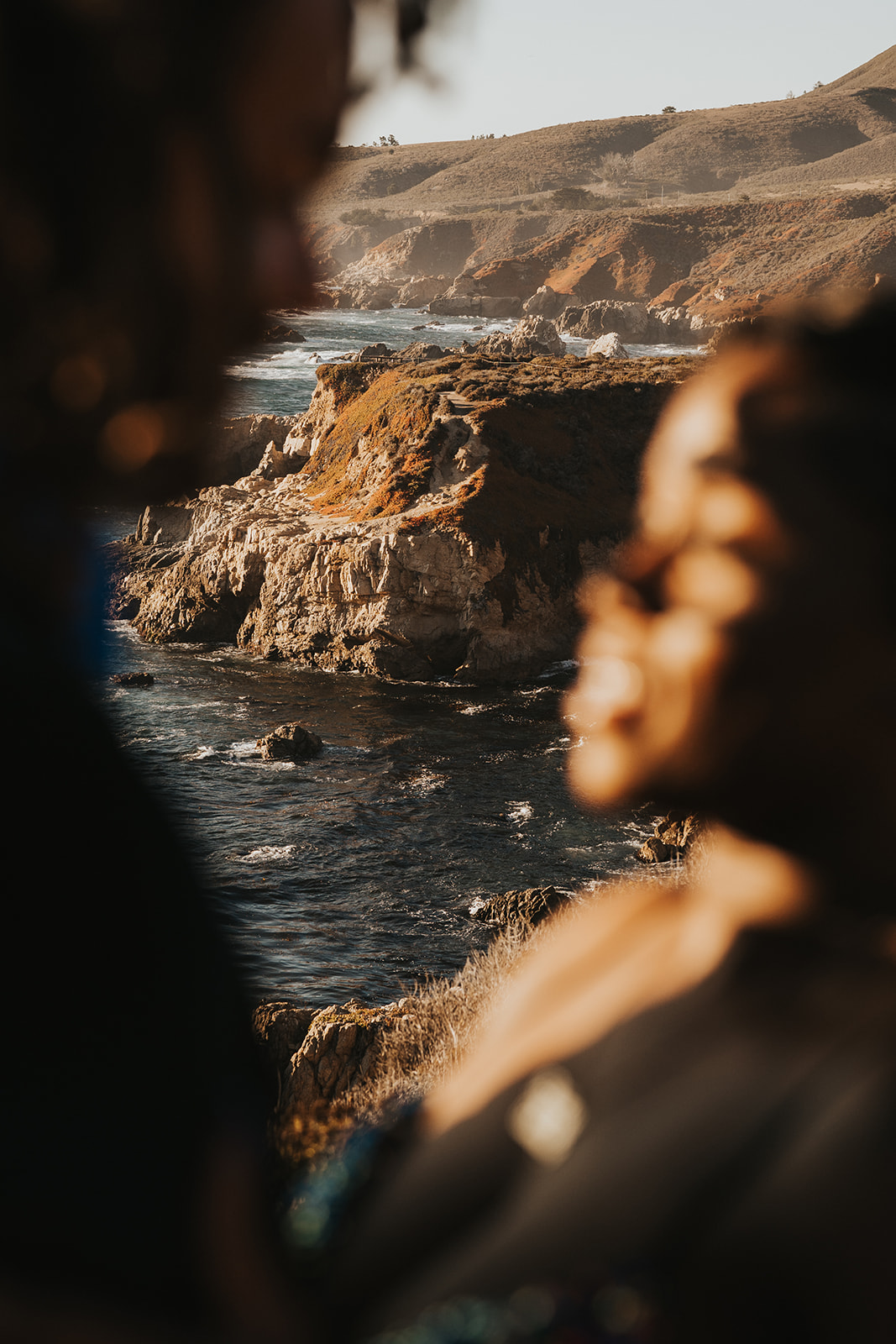 Couple in foreground of image while the Big Sur coastline is in focus behind them