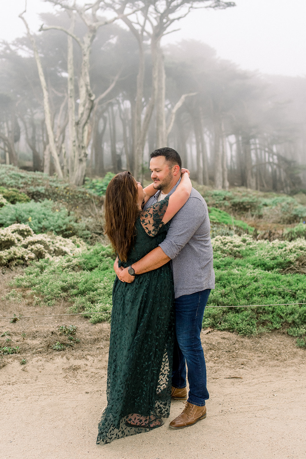 A couple celebrates their engagement in the Cypress trees in fog at Lands End San Francisco CA.