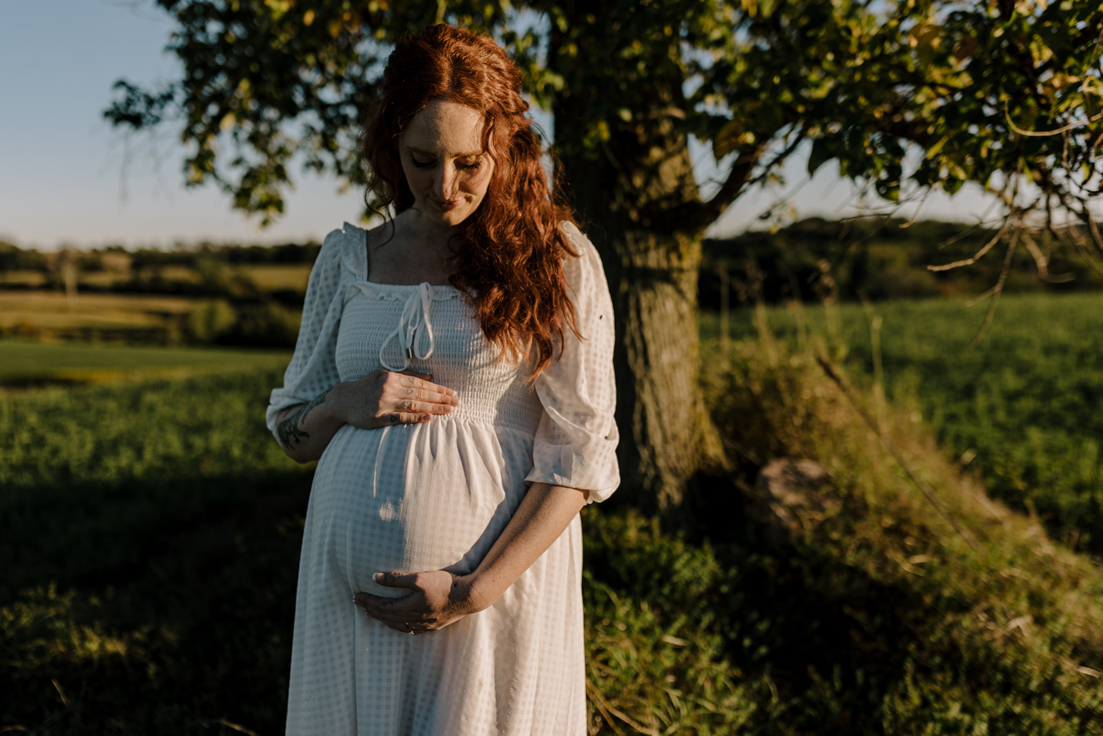 pregnant woman holding belly in white dress in front of tree at sunset