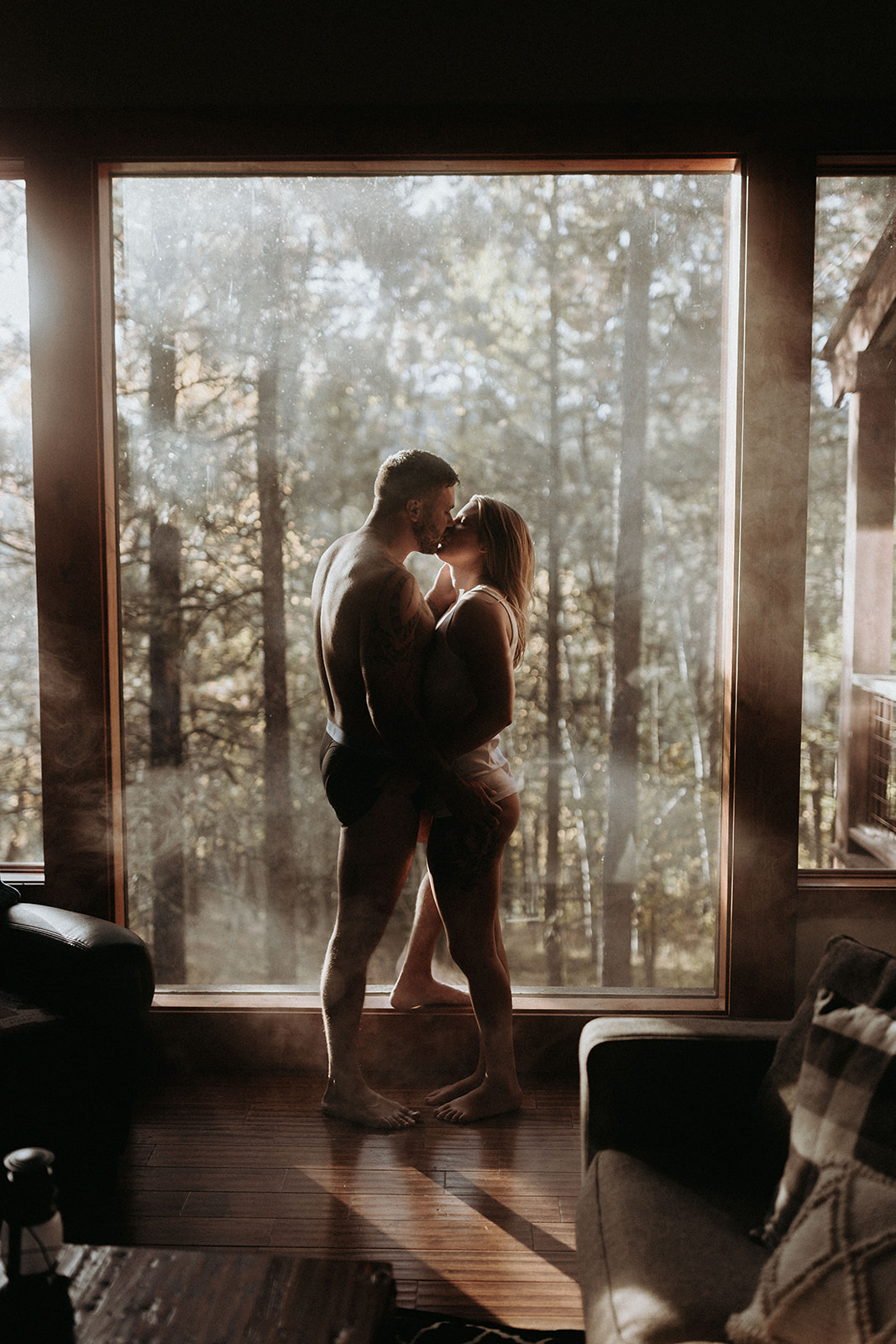A couple kissing in their underwear in their airbnb with the forest outside the window in the background.