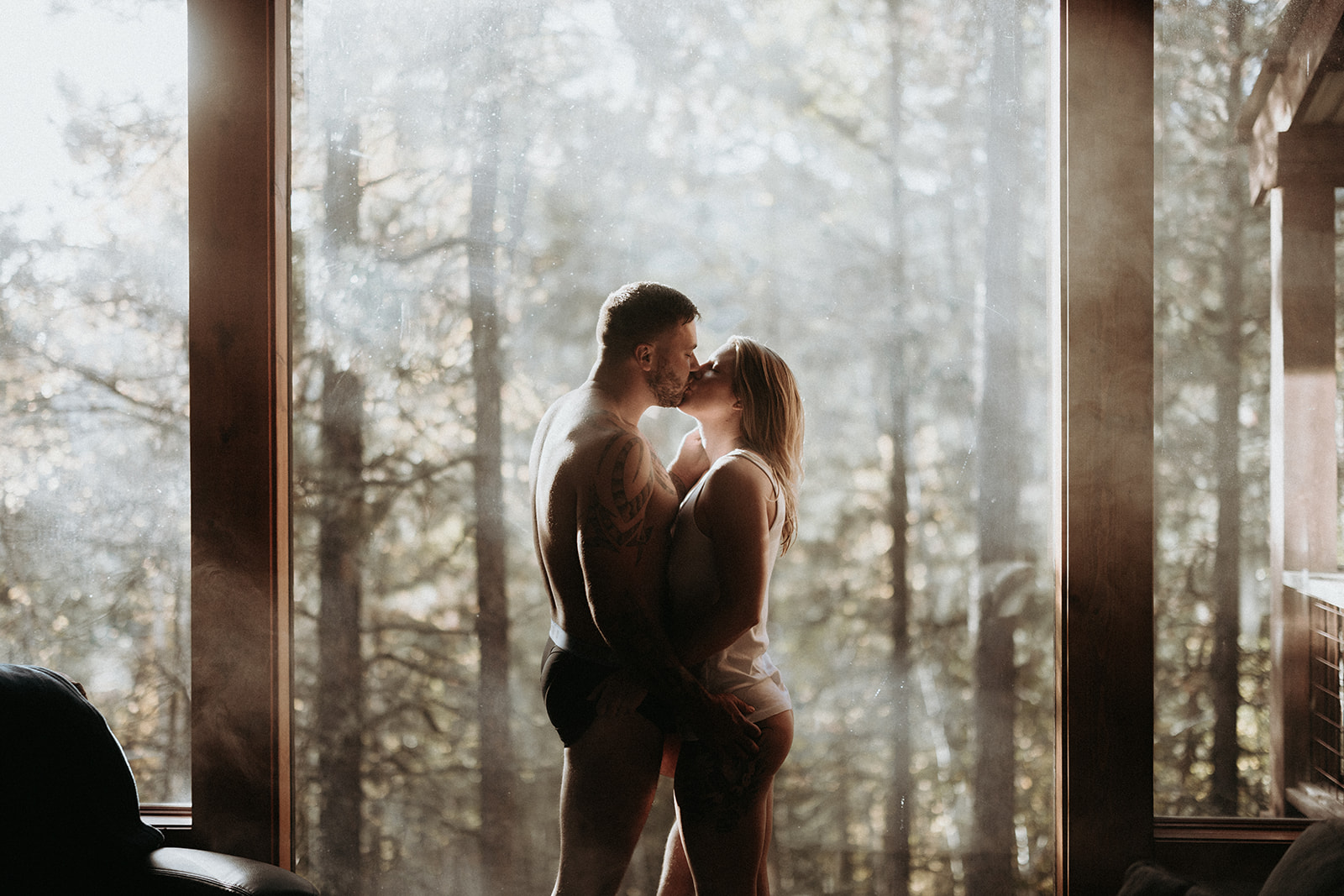 A couple kissing in their underwear in their airbnb boudoir session with the forest outside the window in the background