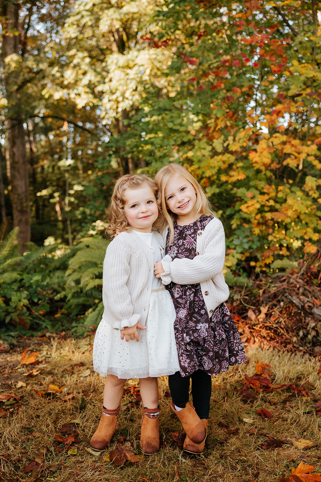 A photo of two young girls hugging each other and smiling at the camera in front of colorful fall foliage.