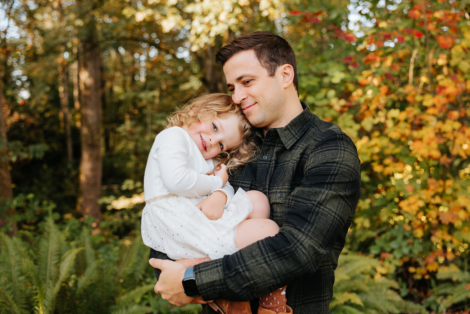 A close-up candid photo of a father holding his daughter in his arms in front of colorful fall foliage in Seattle.