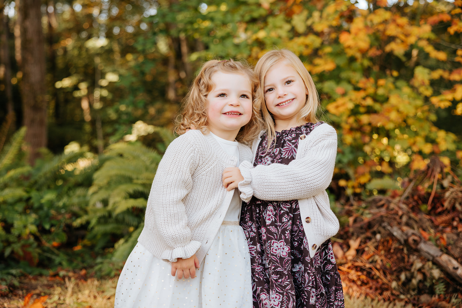 A close-up photo of two young girls hugging each other and smiling at the camera in front of colorful fall foliage.