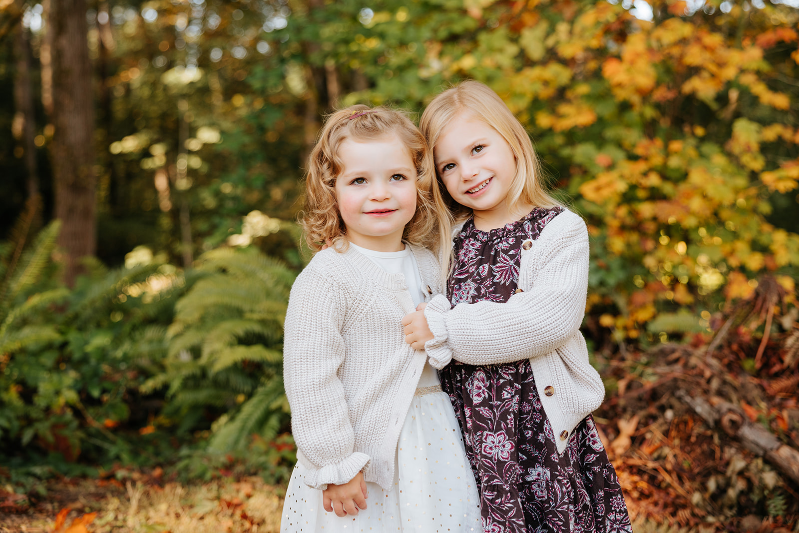 A close-up photo of two young girls hugging each other and smiling at the camera in front of colorful fall foliage.
