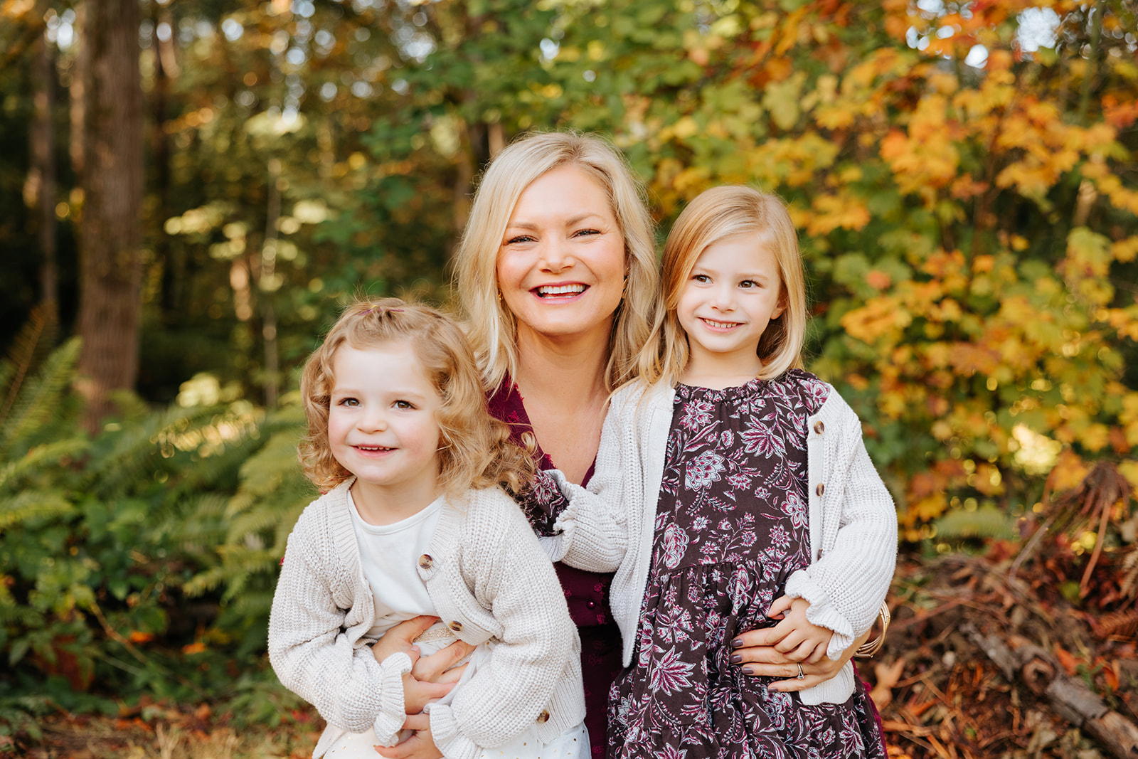 A photo of two young daughters hugging their mother and smiling at the camera in front of colorful fall foliage.