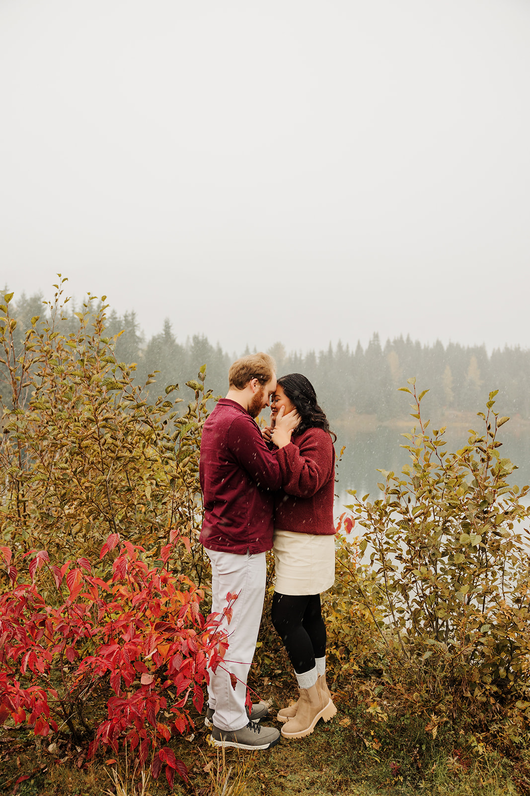 A photo of a loving couple embracing one another near a winter pond in the mountains of Washington while the snow falls.