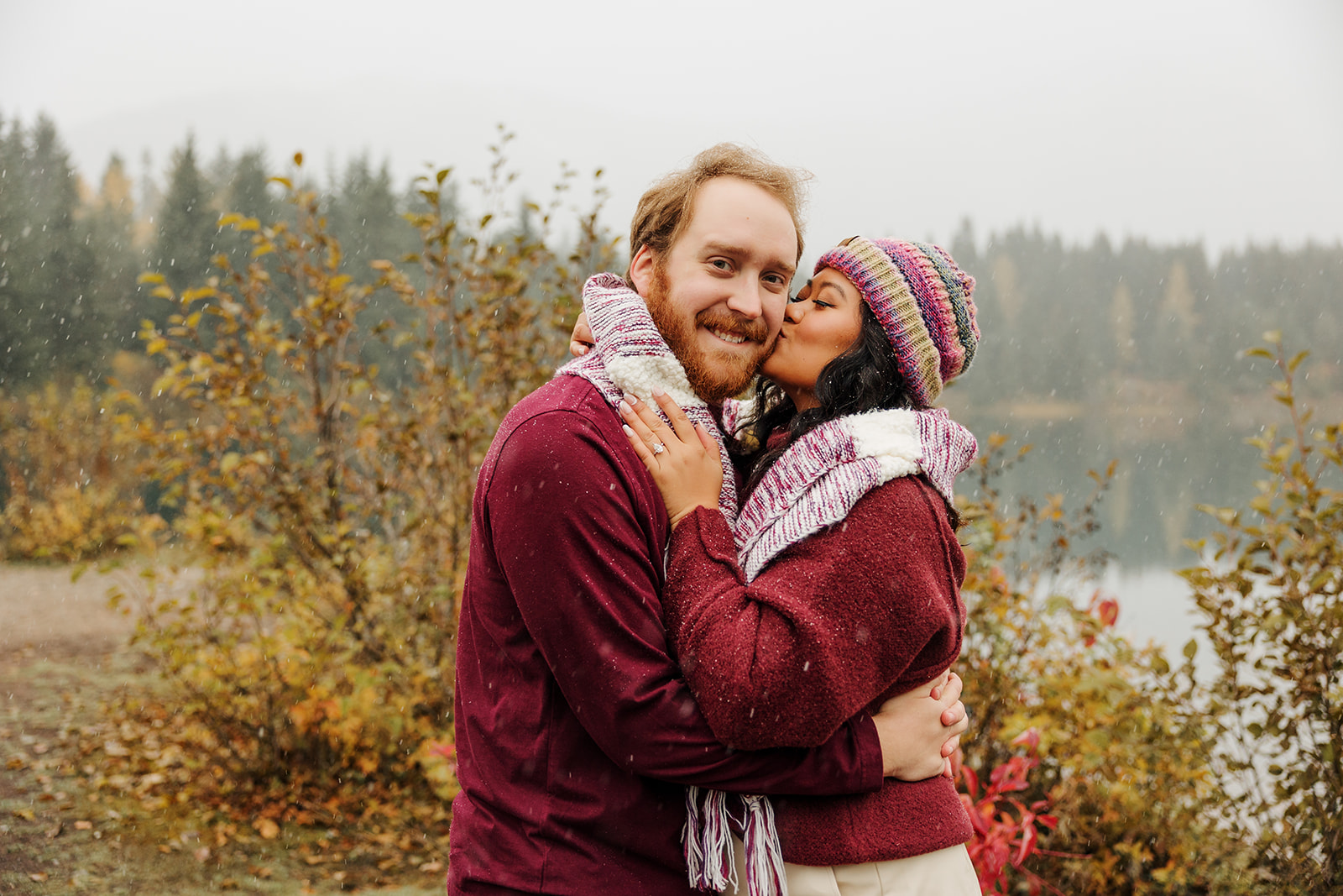 A photo of a loving couple embracing one another near a winter pond in the mountains of Washington while the snow falls.