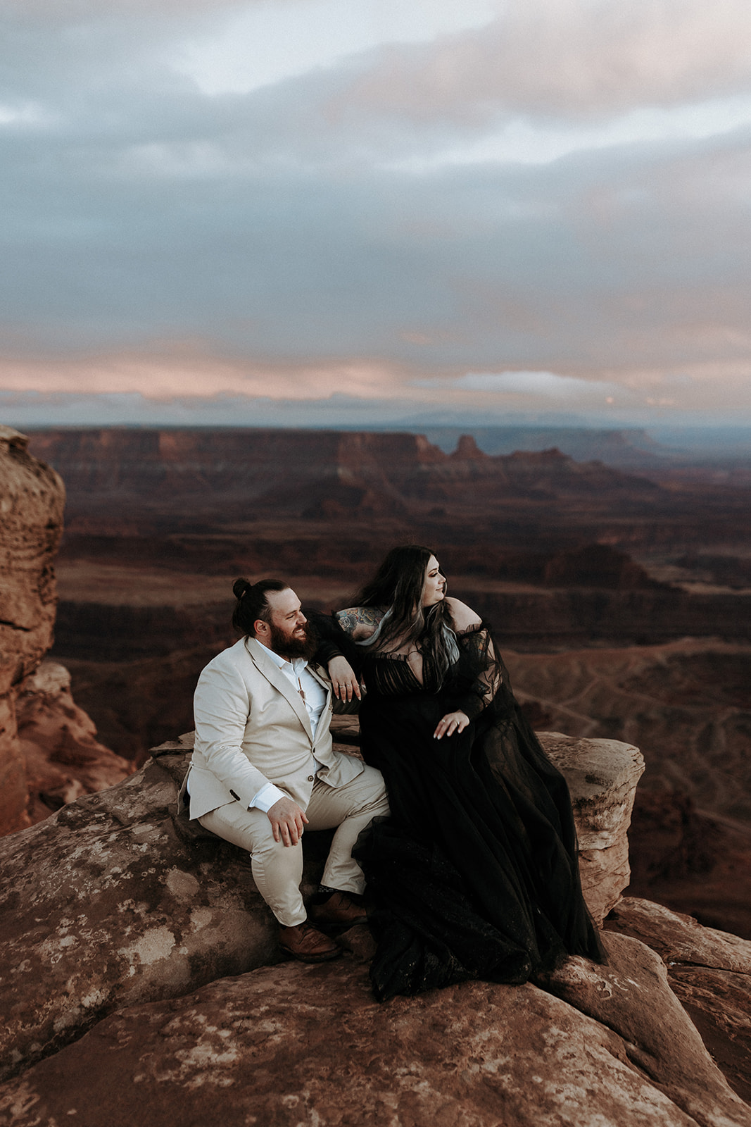 A bride in a black dress and her husband sitting down overlooking a canyon at Dead Horse Point at sunset.