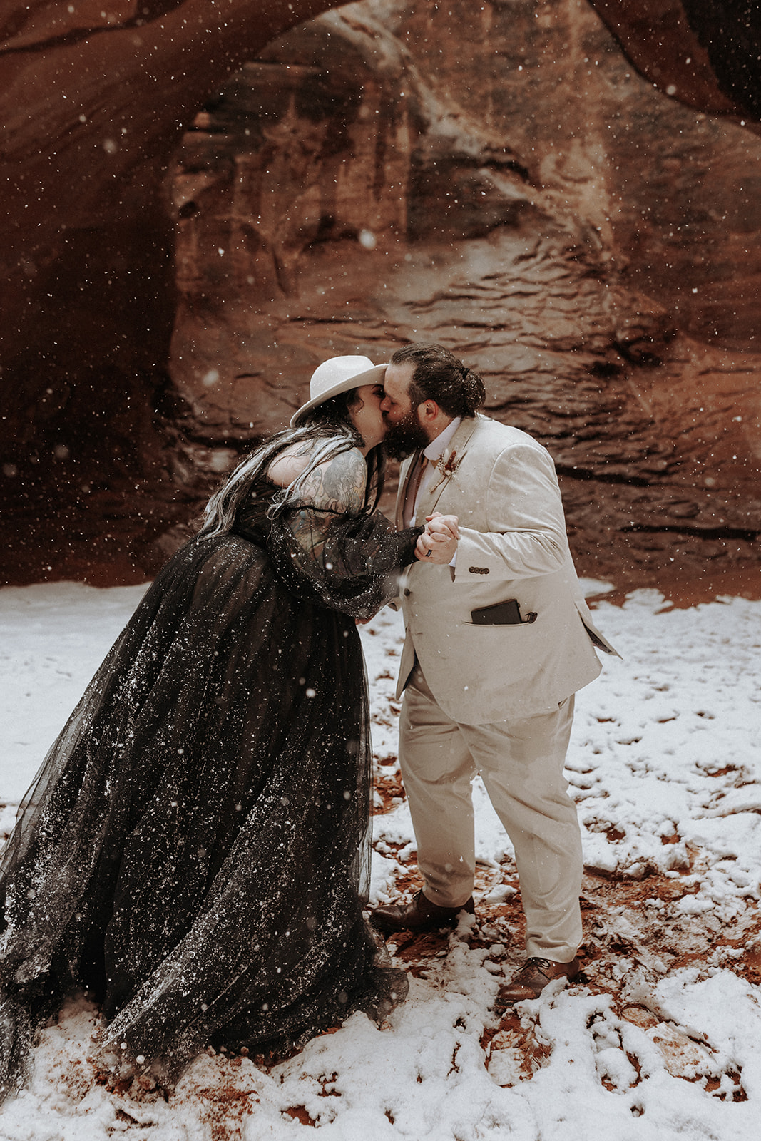 A bride wearing a black dress kissing her husband at Sand Dune Arch in Arches National Park in the snow.
