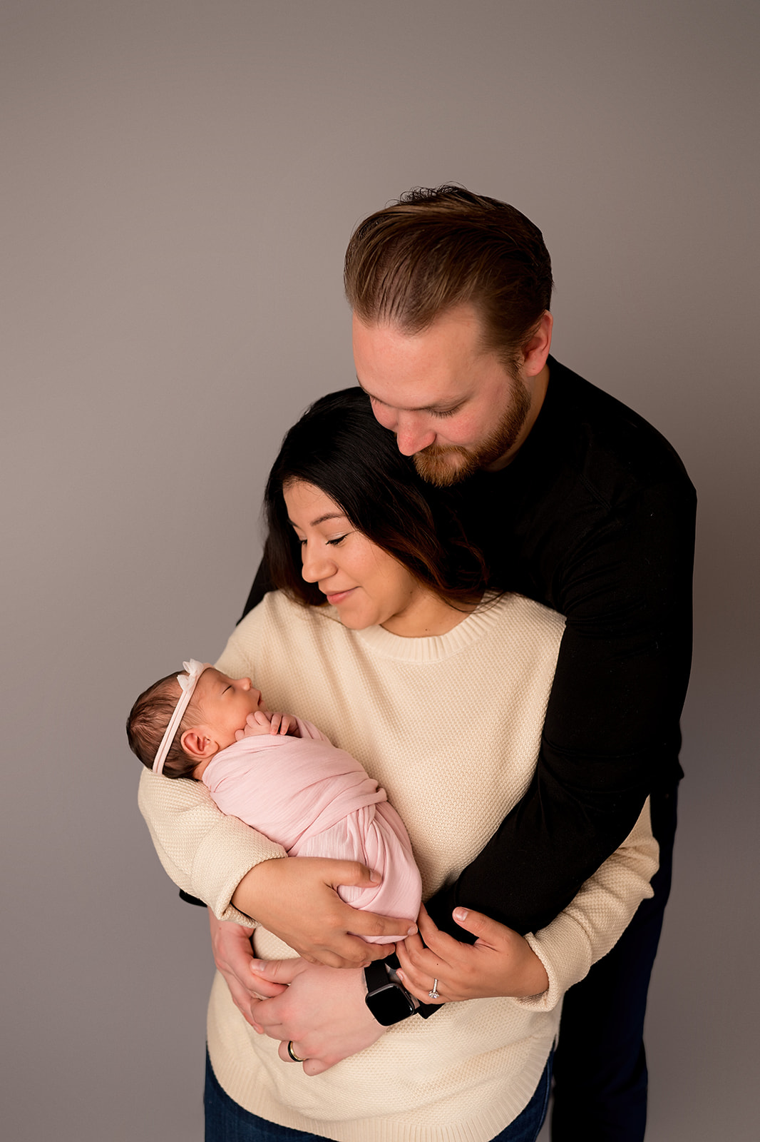 First time parents with arms wrapped around each other holding newborn baby girl swaddled in pink with delicate headband