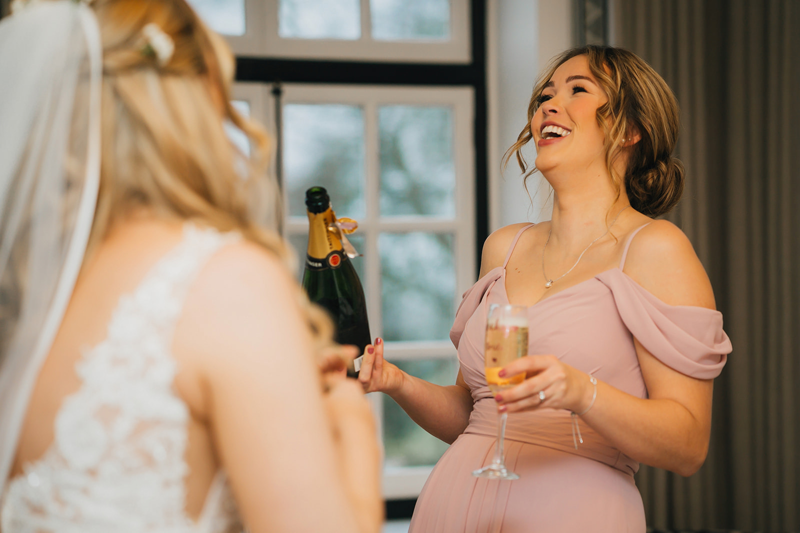 a bridesmaid and the bride laugh, champagne glasses in hand
