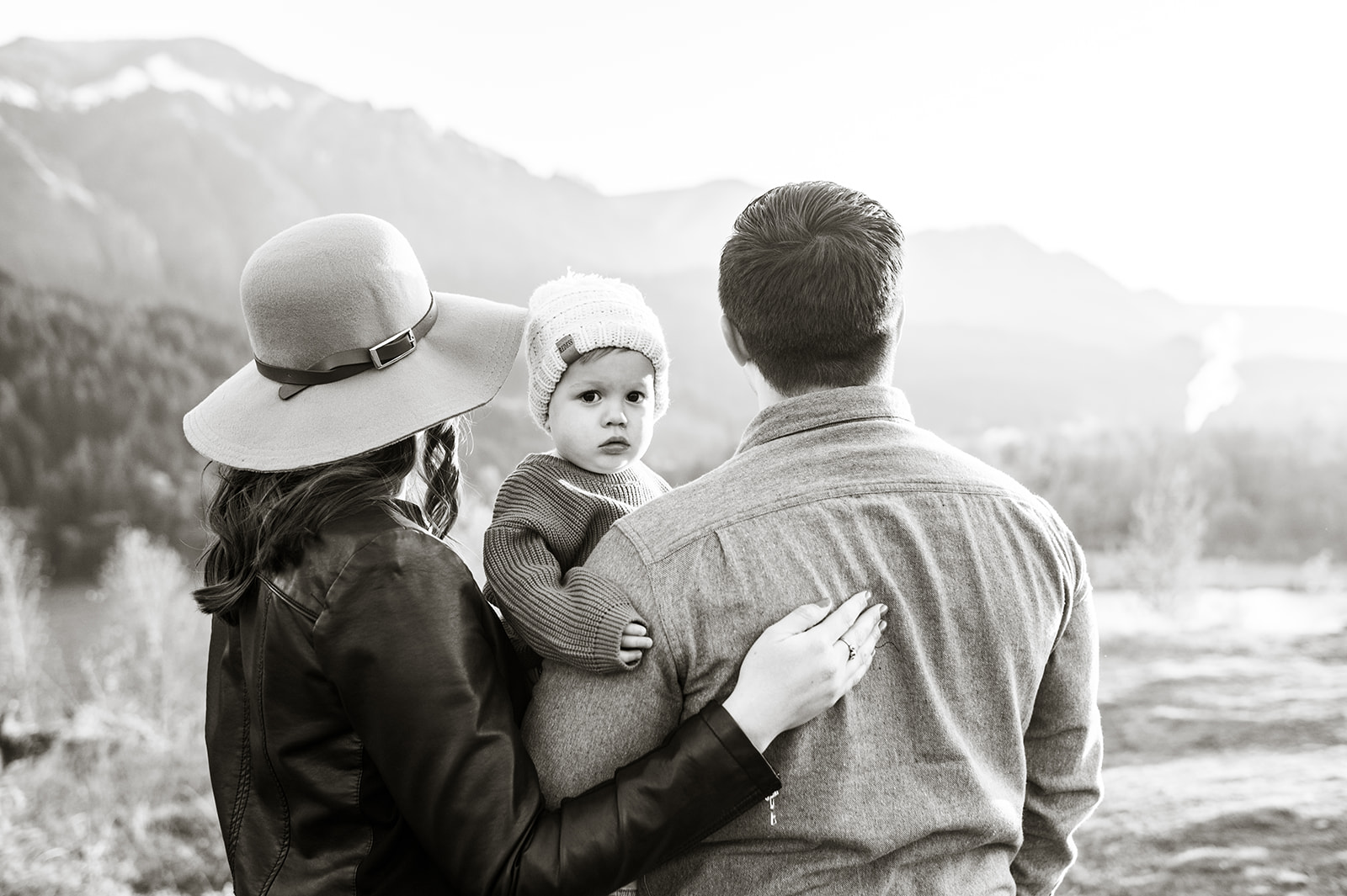 a black and white image of a family of three stand in front of mountains at sunset