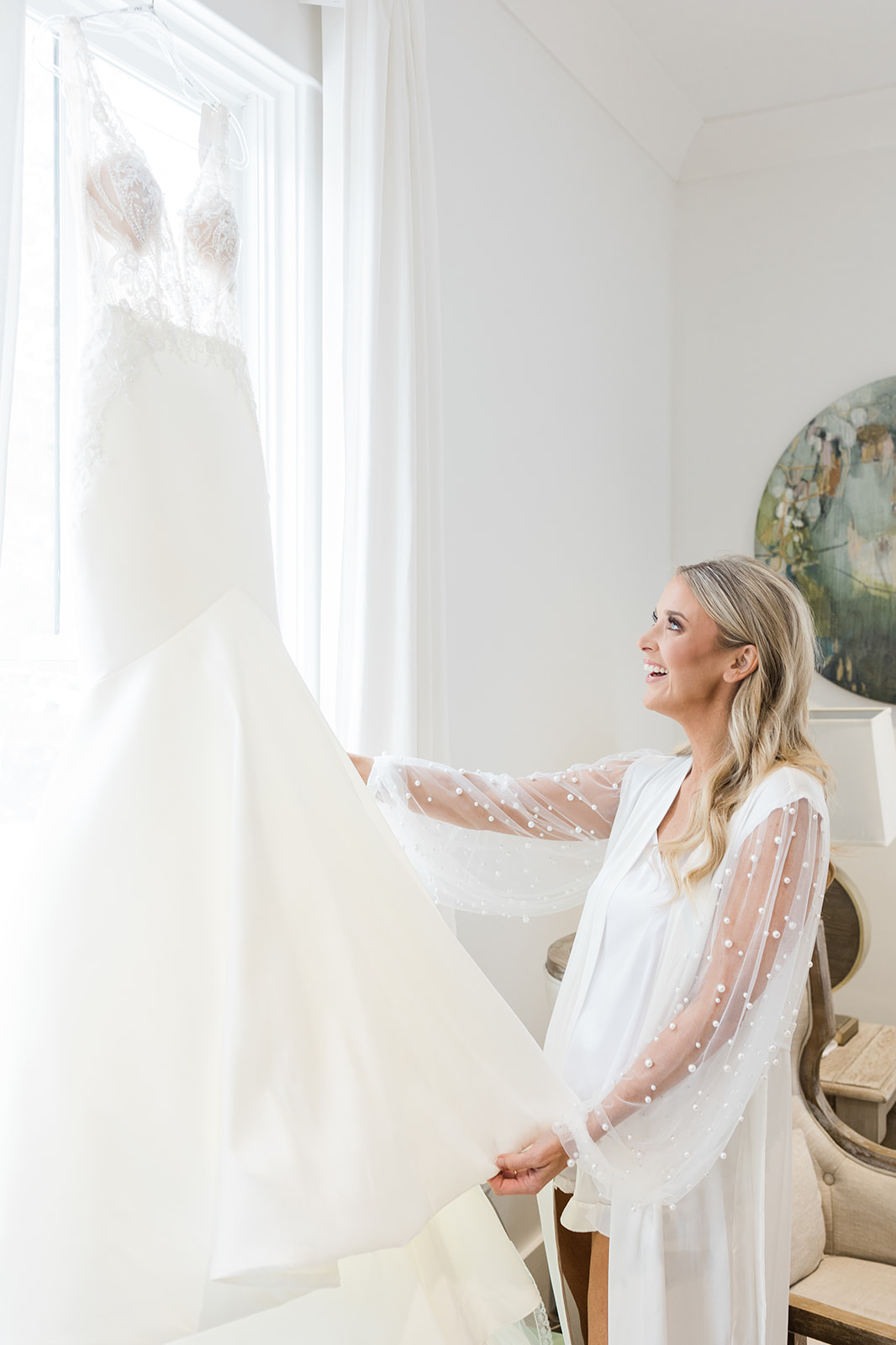 A blushing bride looks excitedly at her wedding gown in the glow of the natural sunlight through her bridal suite window