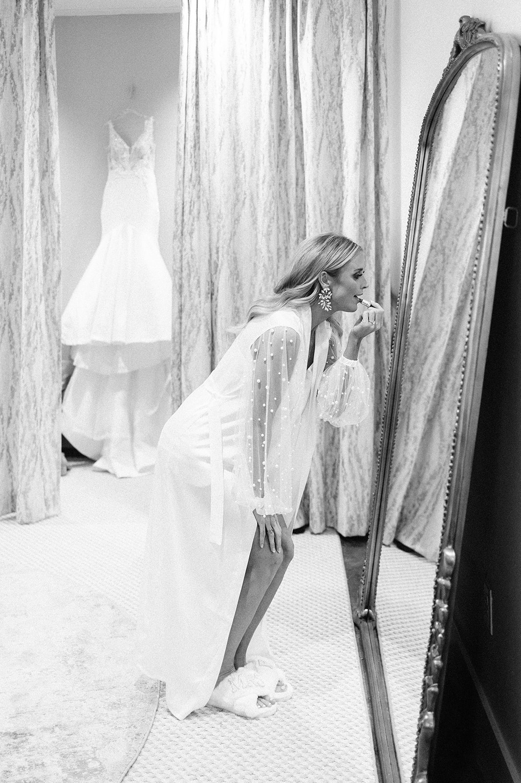 A black&white image where abride puts on her makeup while looking at herself in the mirror with her wedding gown 