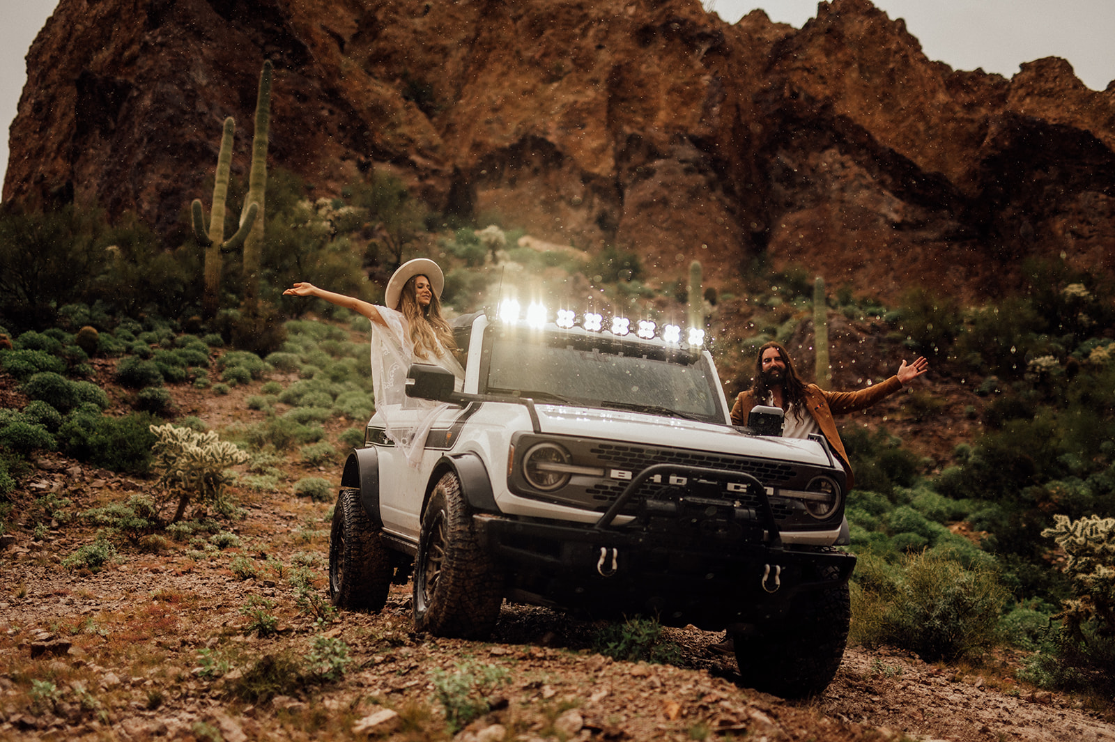 Elopement couple leaning out the windows of a Ford Bronco with rain coming down in front of mountains and saguaro cactus